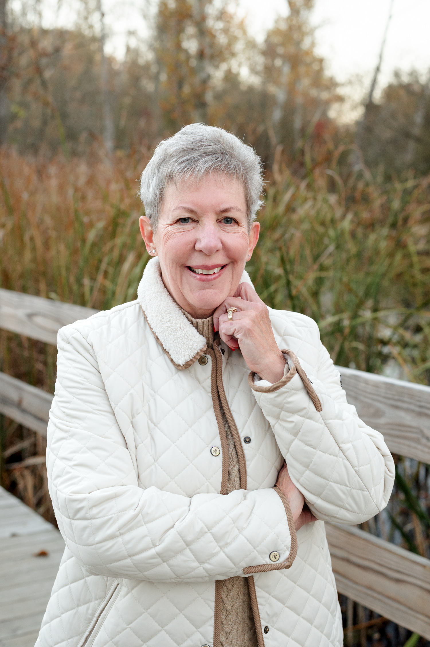 Person wearing a white quilted jacket and tan scarf poses outdoors on a wooden deck during autumn.