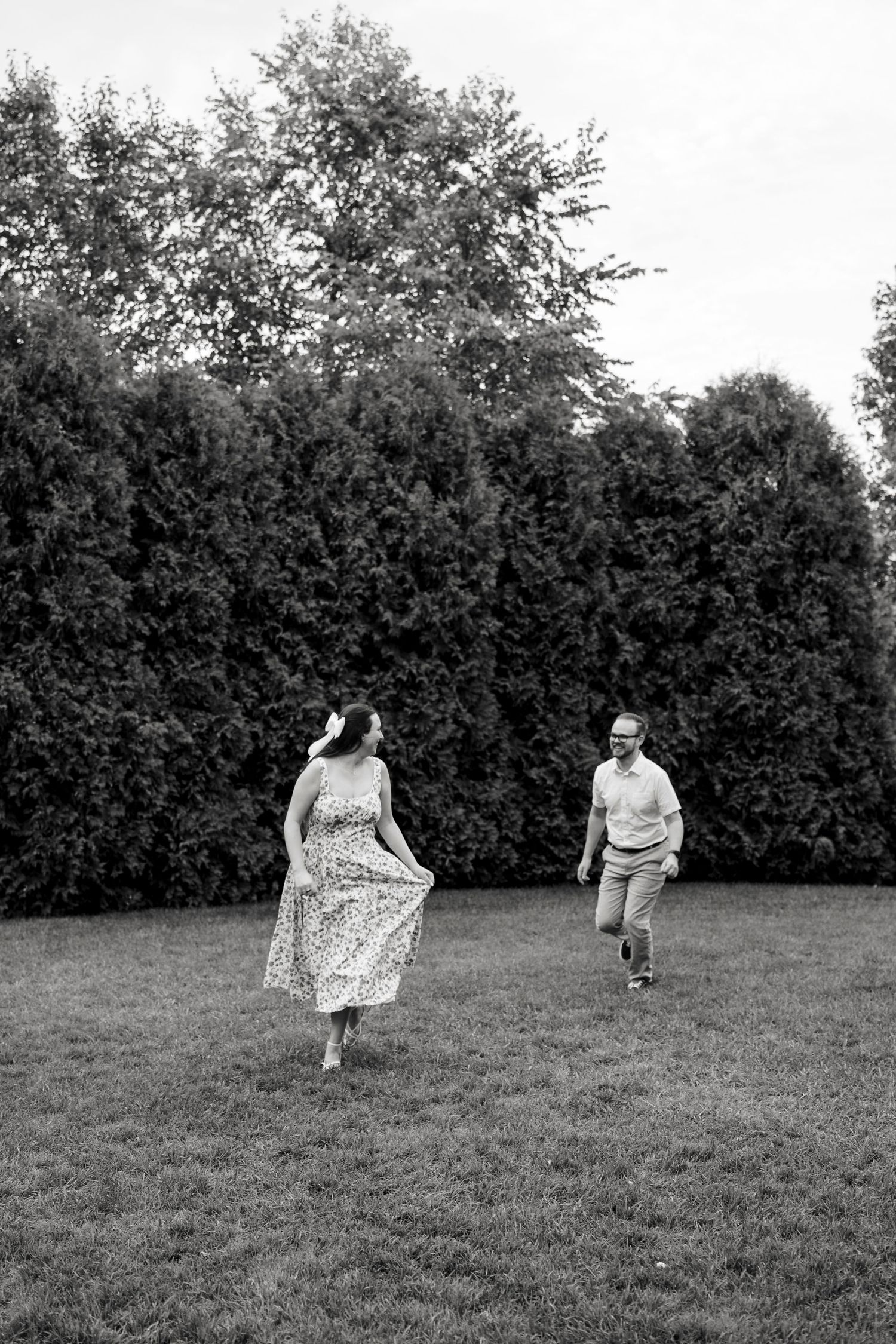 Black and white photo of a couple running and playing joyfully on a green lawn with tall hedges in the background.