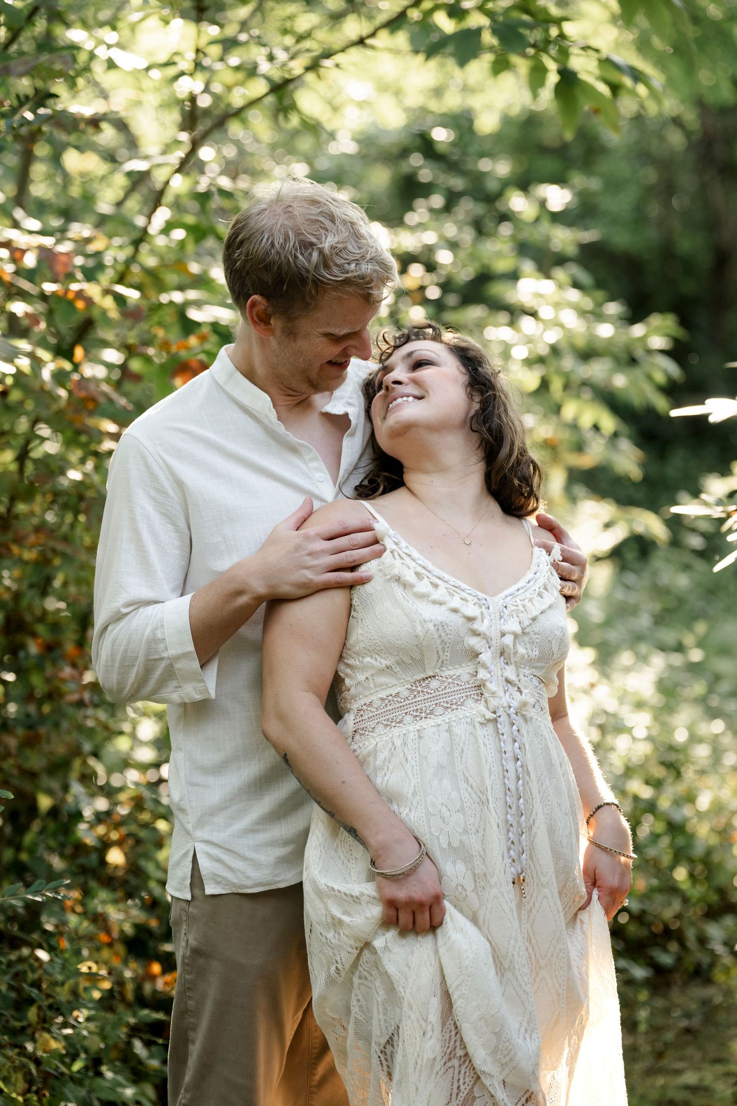 Couple embracing in lush green forest, woman in white lace dress laughing joyfully as man holds her close.
