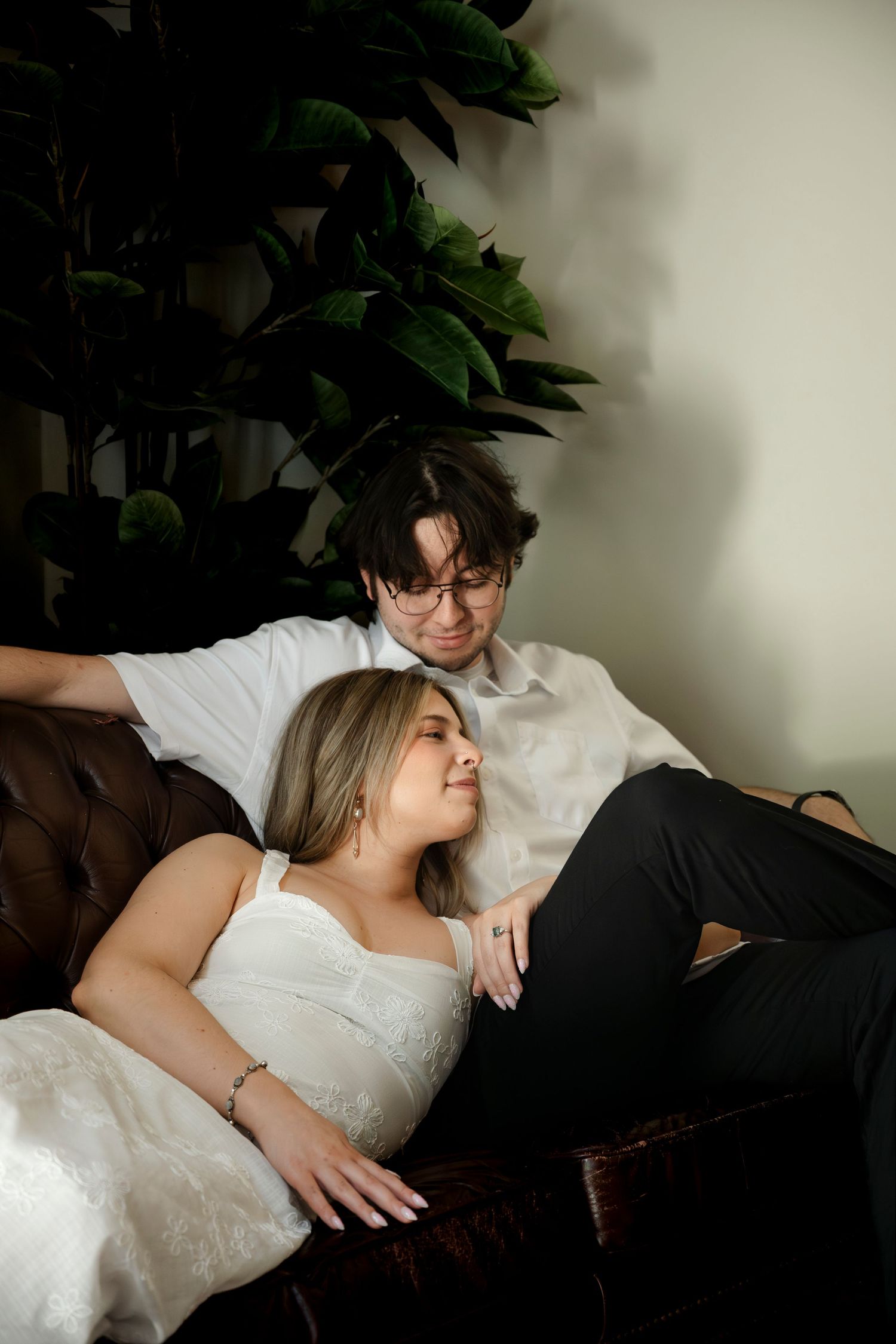 Couple relaxing on leather sofa, woman in white dress leaning back on man in white shirt, with green plant backdrop.