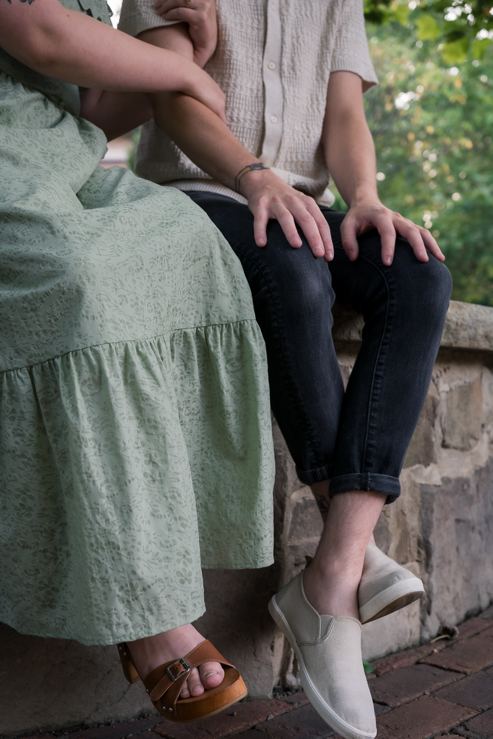 Two people sitting on a stone wall outdoors, one in a green dress with sandals, the other in dark jeans and beige slip-on shoes.