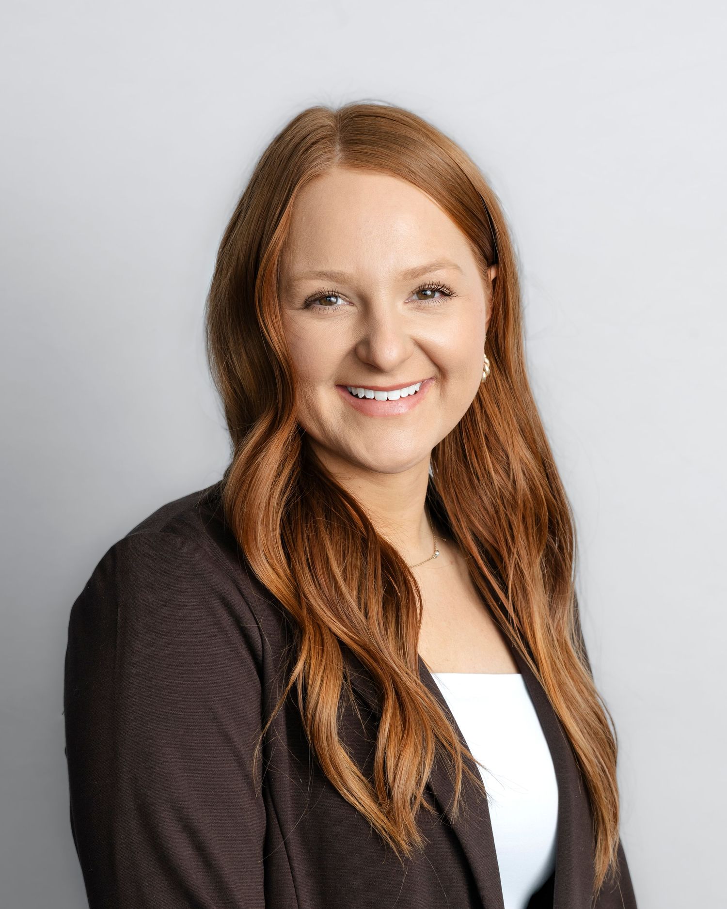 Professional headshot of a person with long auburn hair wearing a dark blazer and white top against a light gray background.