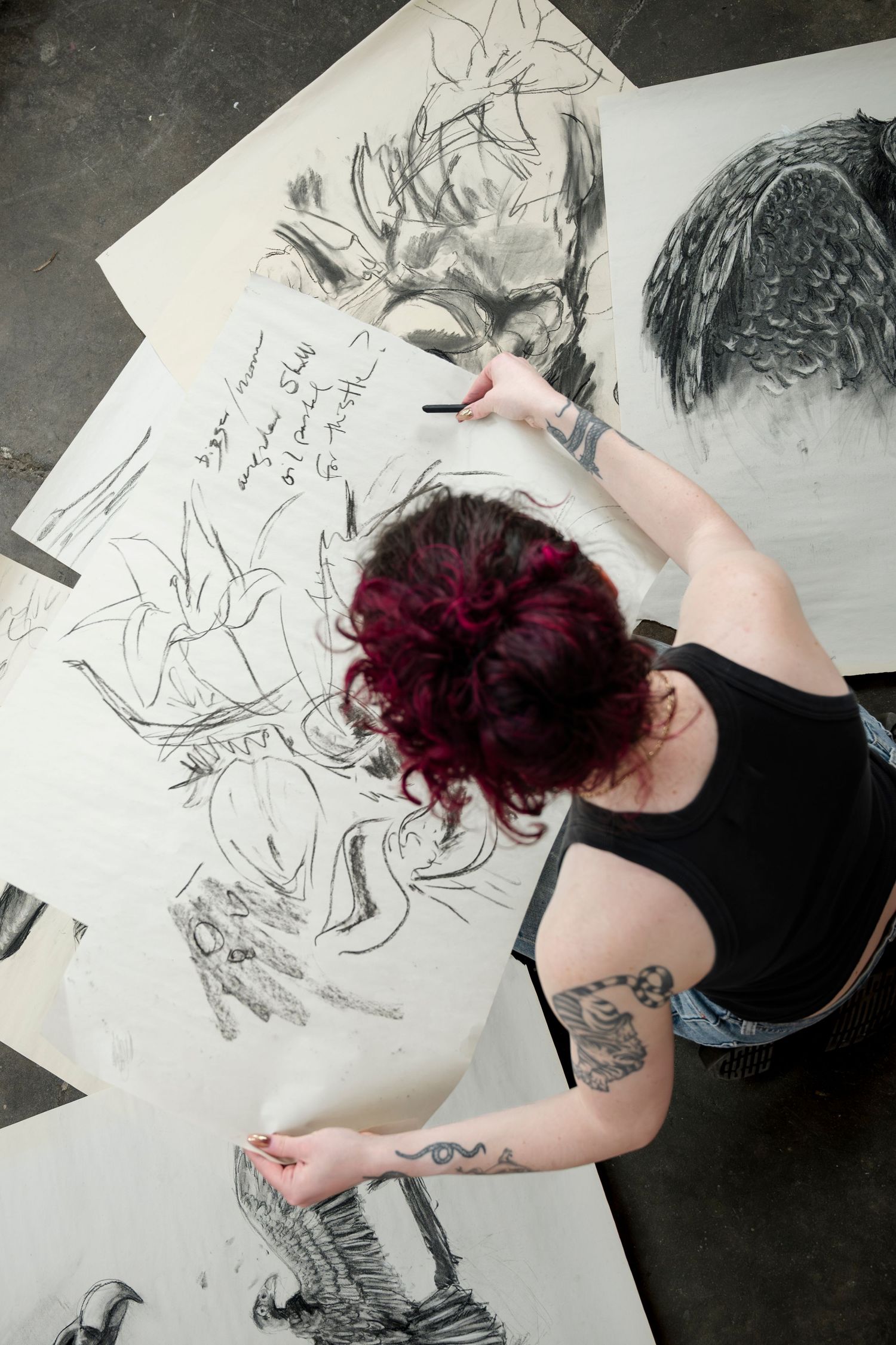 Artist working on large-scale charcoal drawing while sitting on floor in studio space.