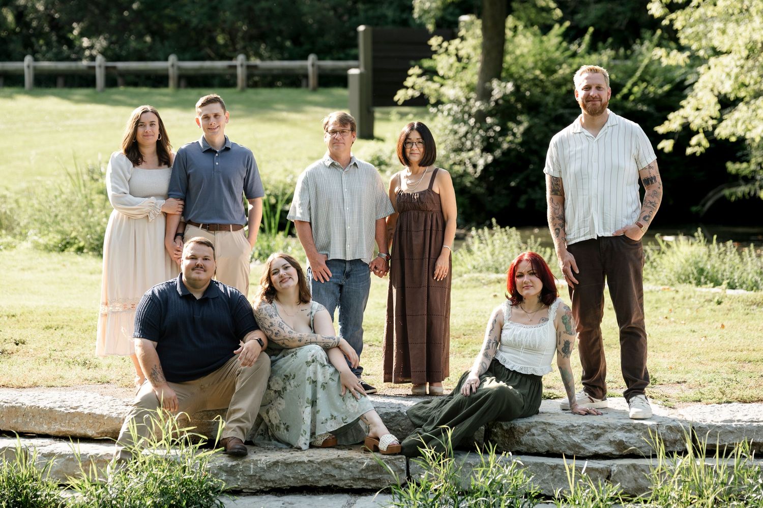 A large family of eight poses outdoors in a lush green park setting for a casual portrait photo.