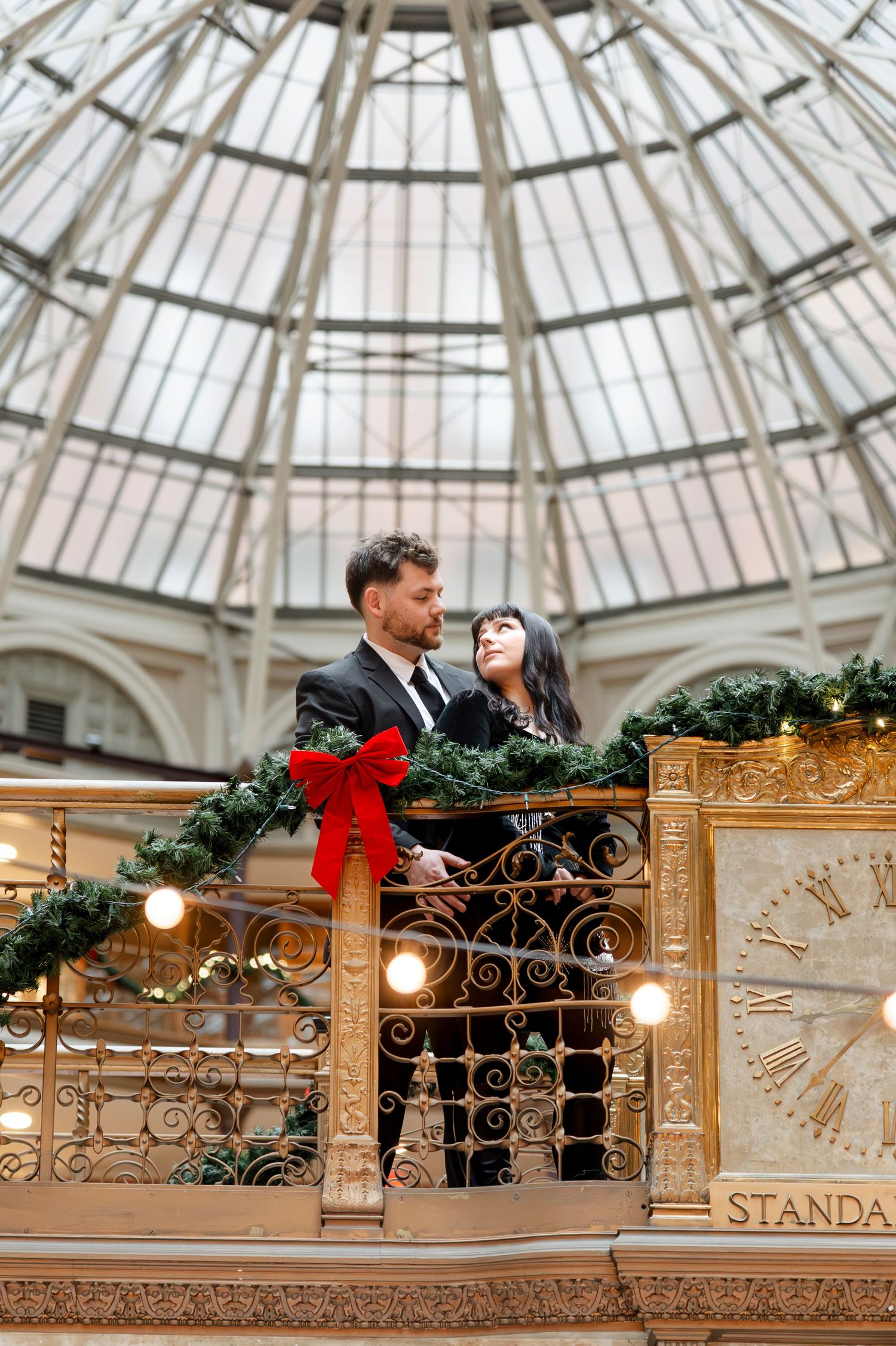 Couple poses on ornate gold balcony with Christmas garland and red bow under glass dome.