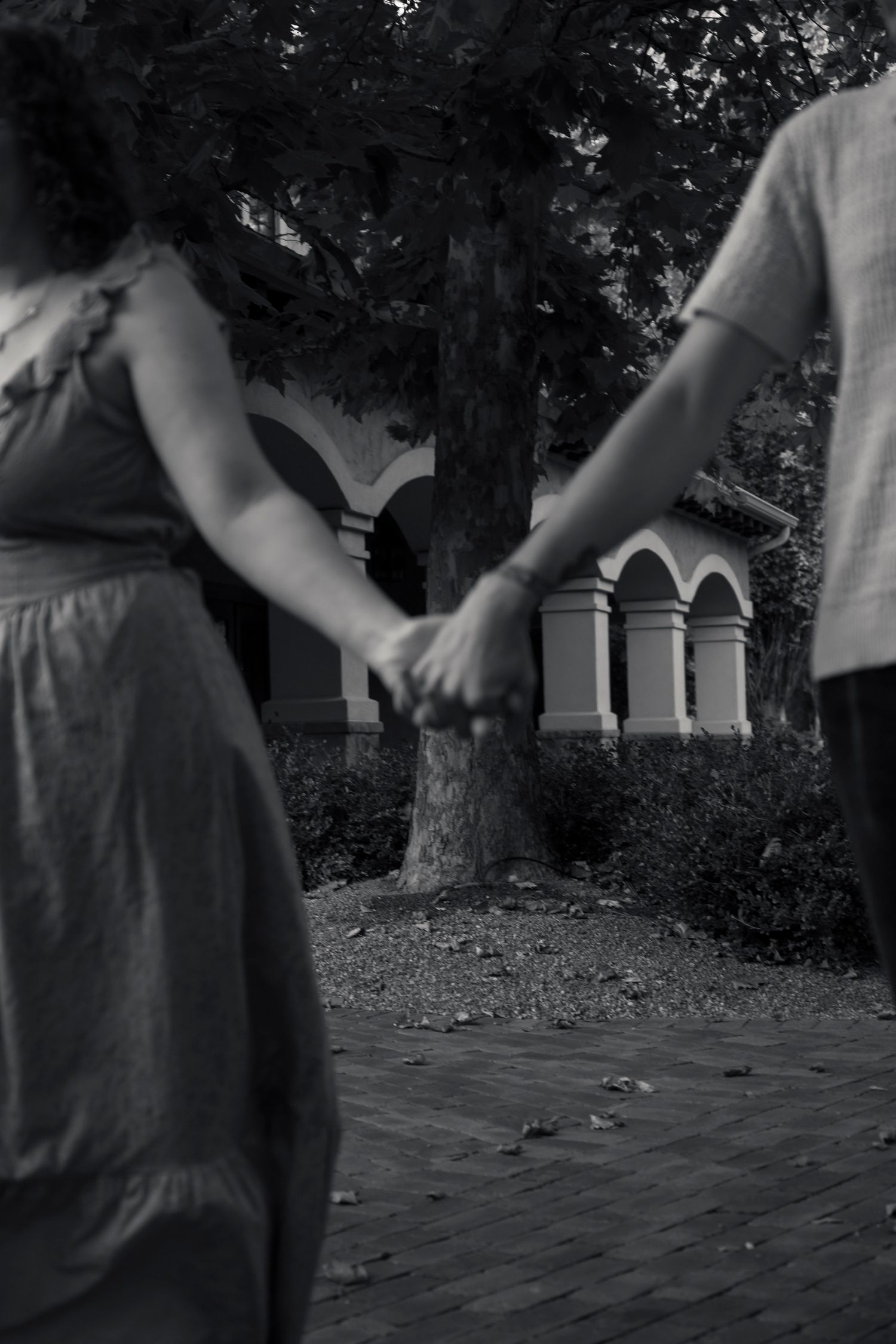Black and white photo of a couple holding hands while walking along a brick path near a historic building.