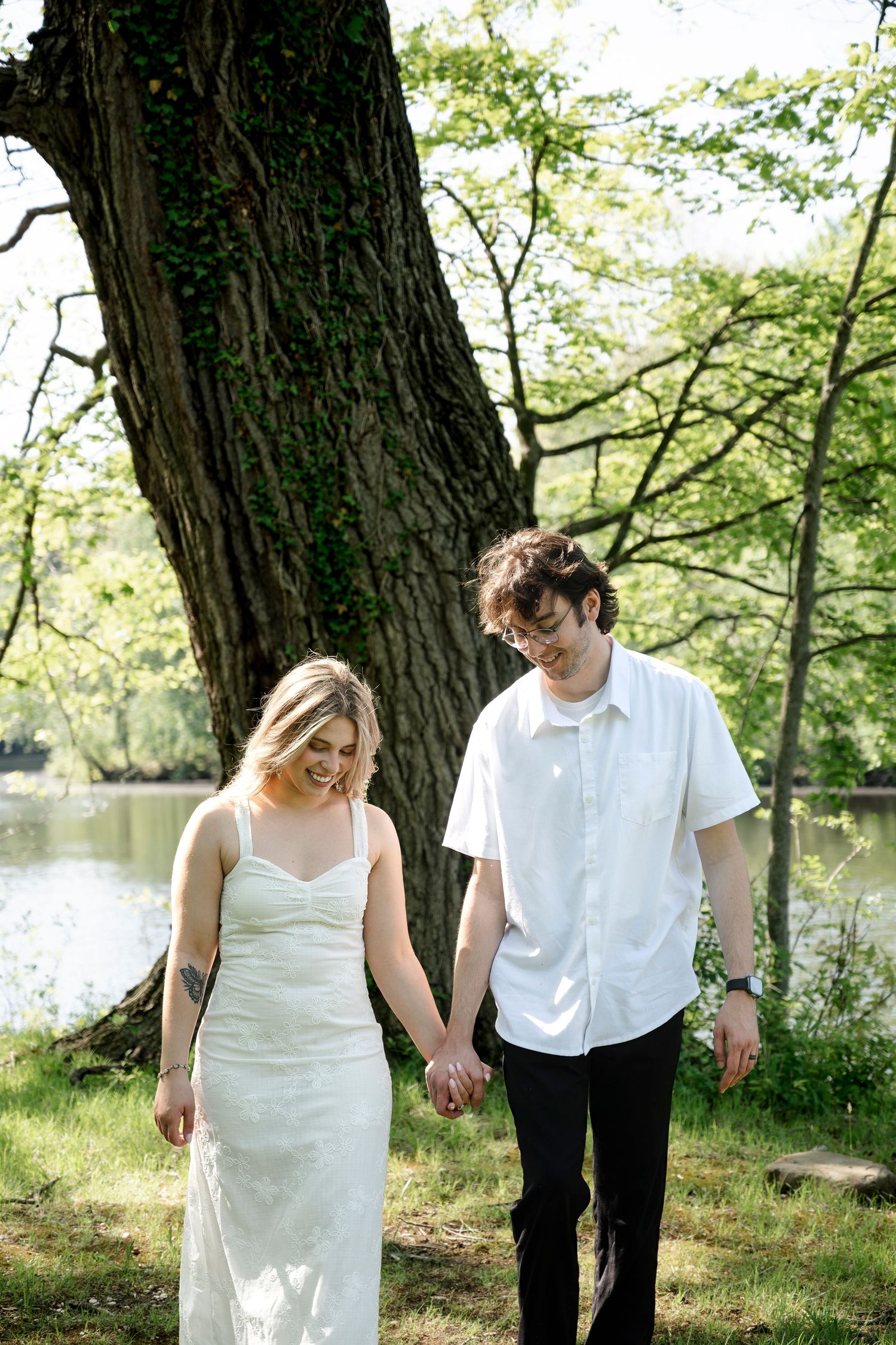 A couple holds hands while walking near a large tree by a pond, the woman in a white dress and man in a white shirt.