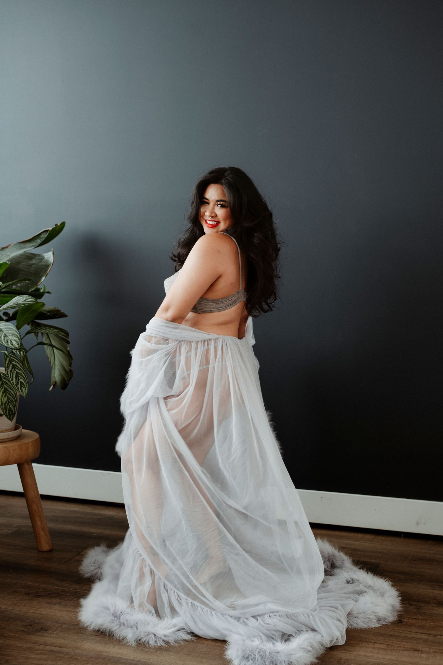 A figure in a flowing white sheer gown poses against a dark gray wall with a houseplant in the corner.