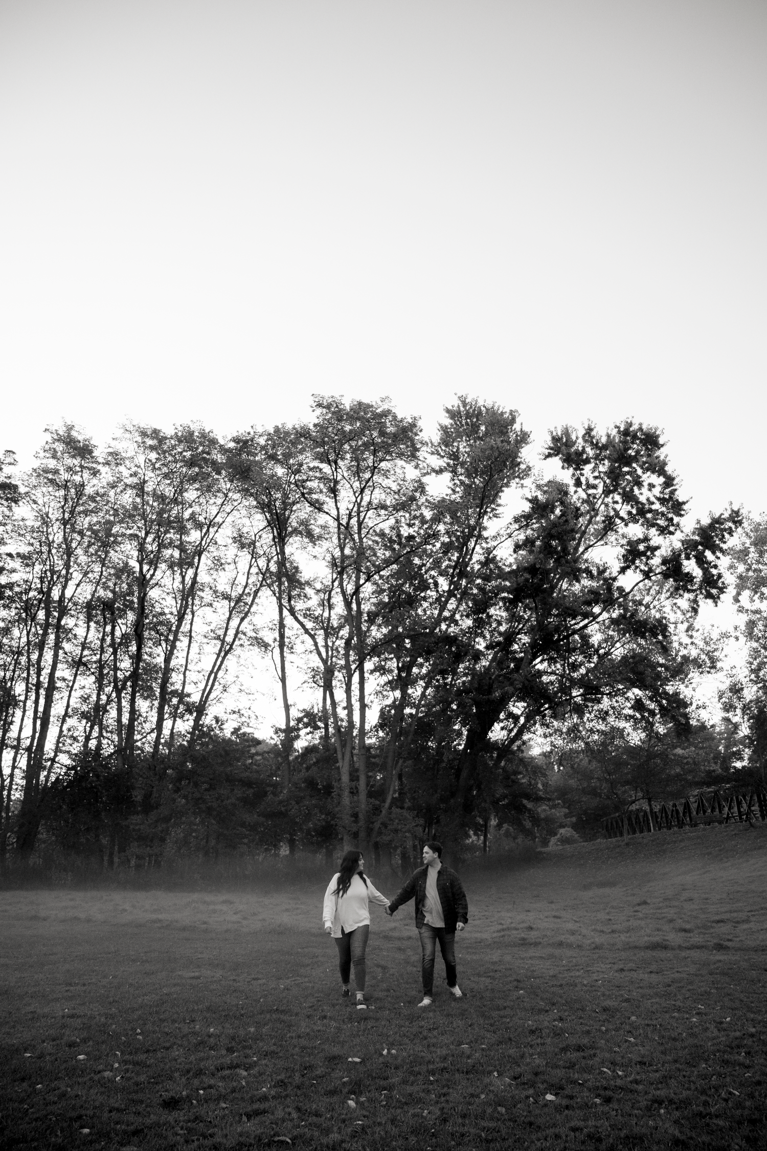 Black and white panoramic sequence shows silhouettes of a couple walking together across an open field with trees in background.