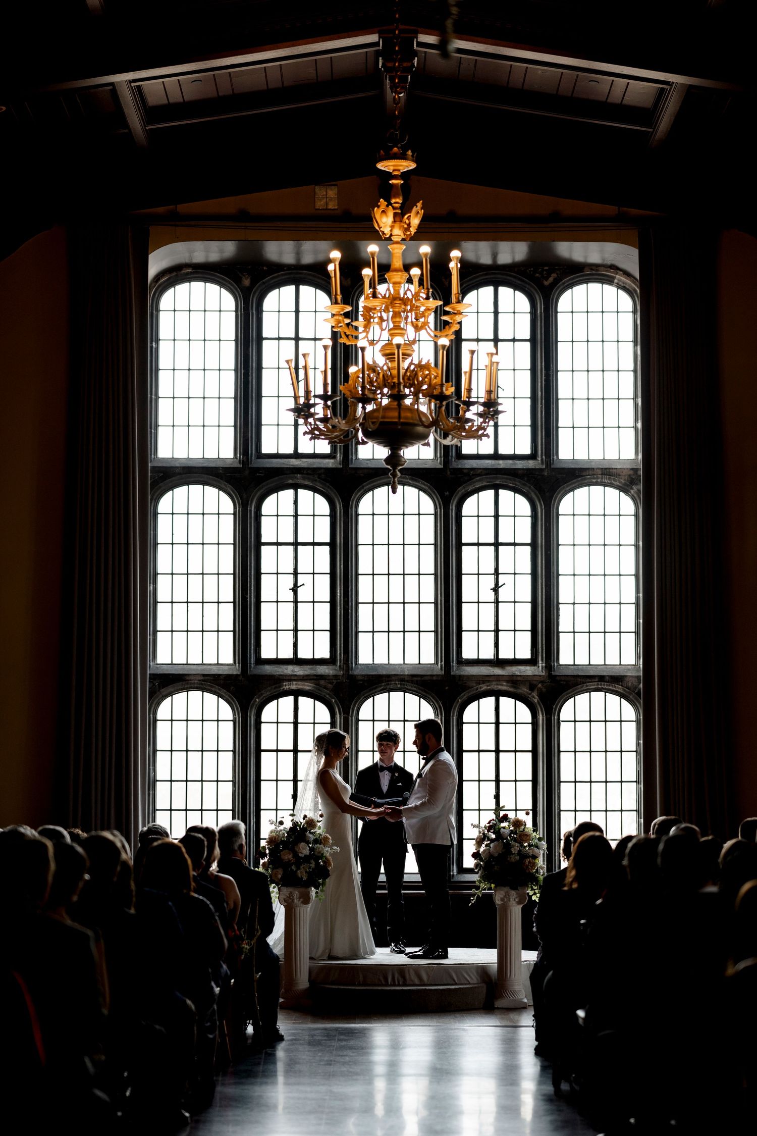 A wedding ceremony taking place in a grand hall with ornate windows and a large chandelier illuminating the scene.