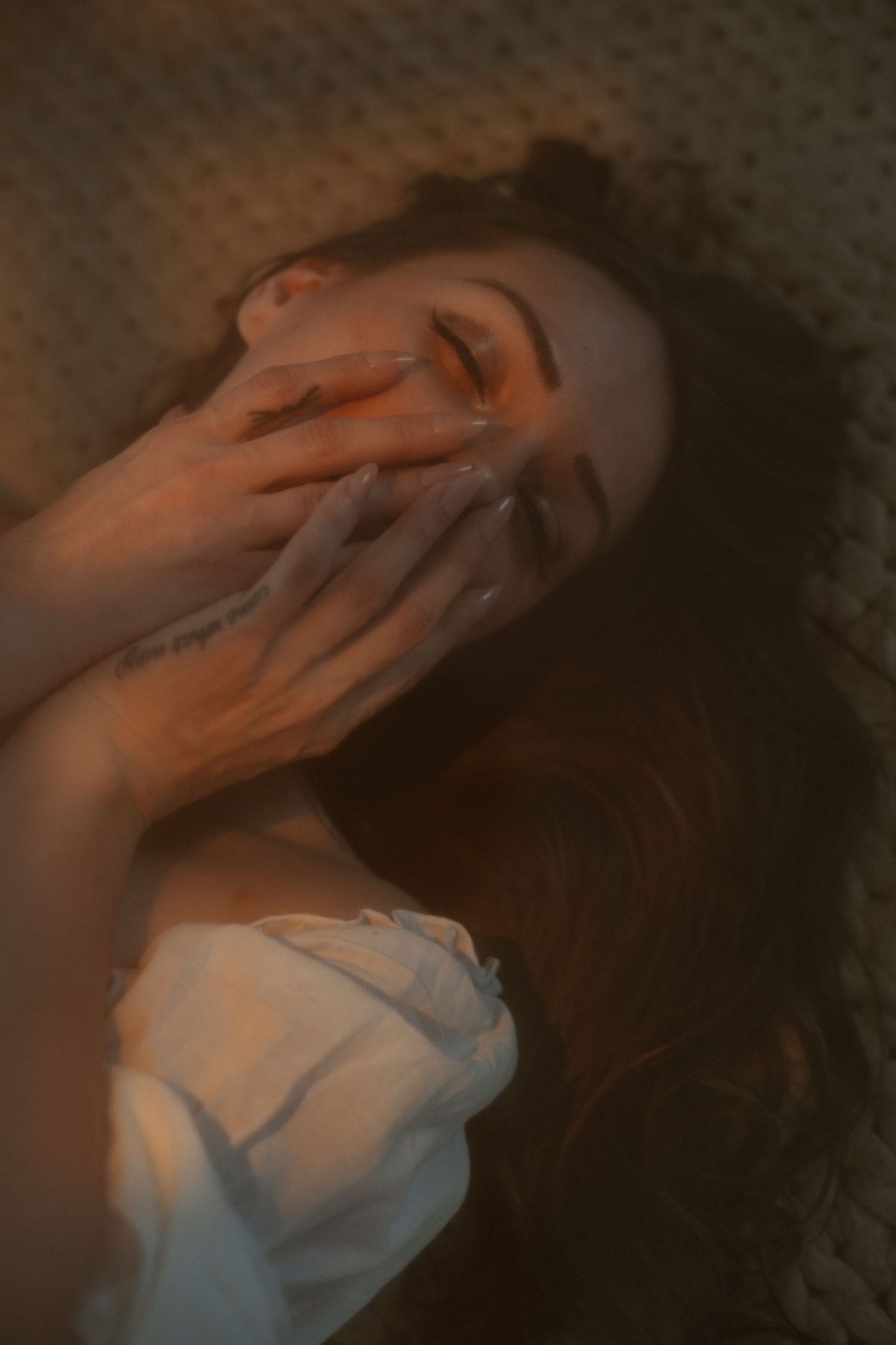 Woman lying down with long hair, hands covering her face, bathed in warm golden light on textured surface.