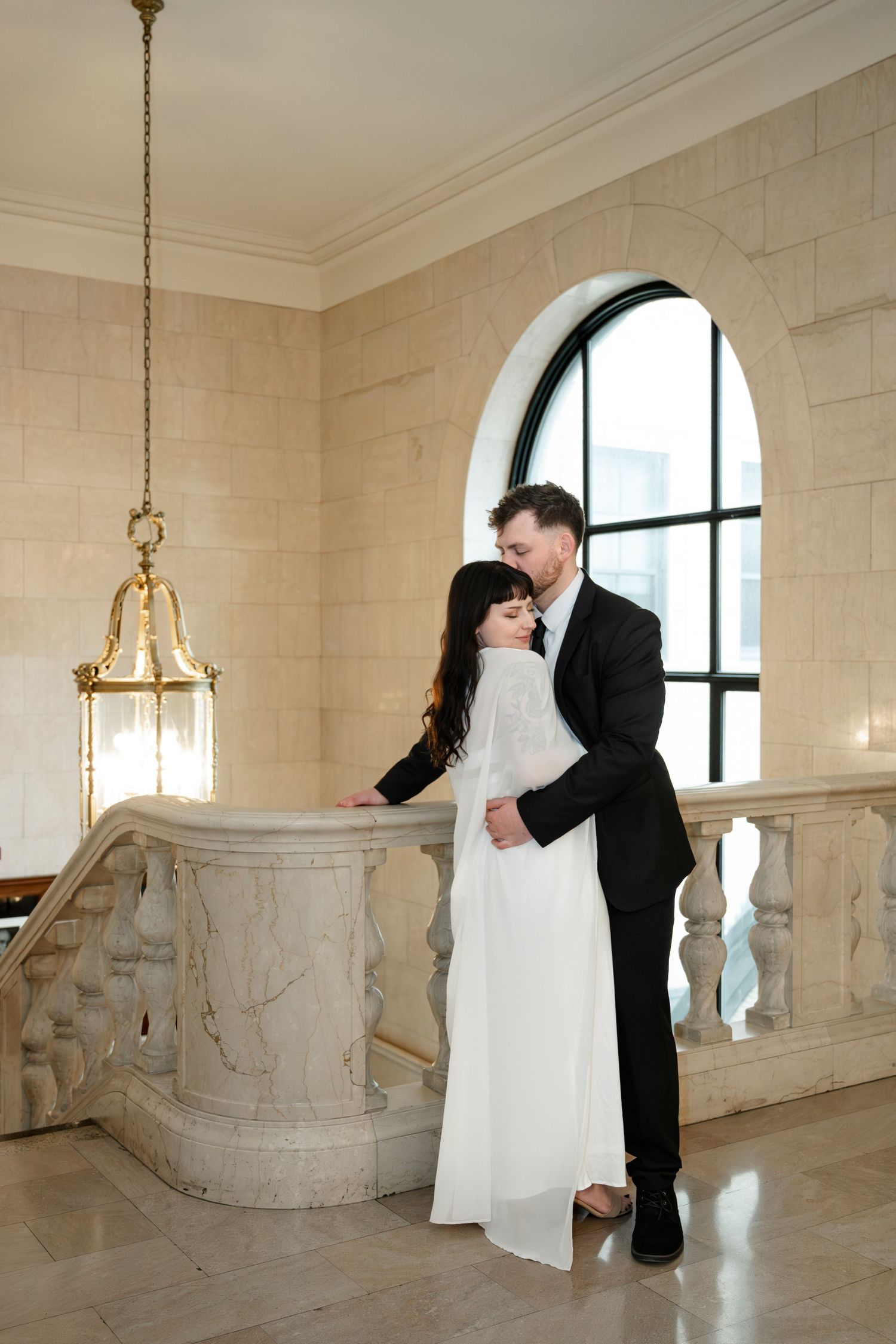 Bride and groom embracing near marble balustrade in elegant beige stone room with arched window.
