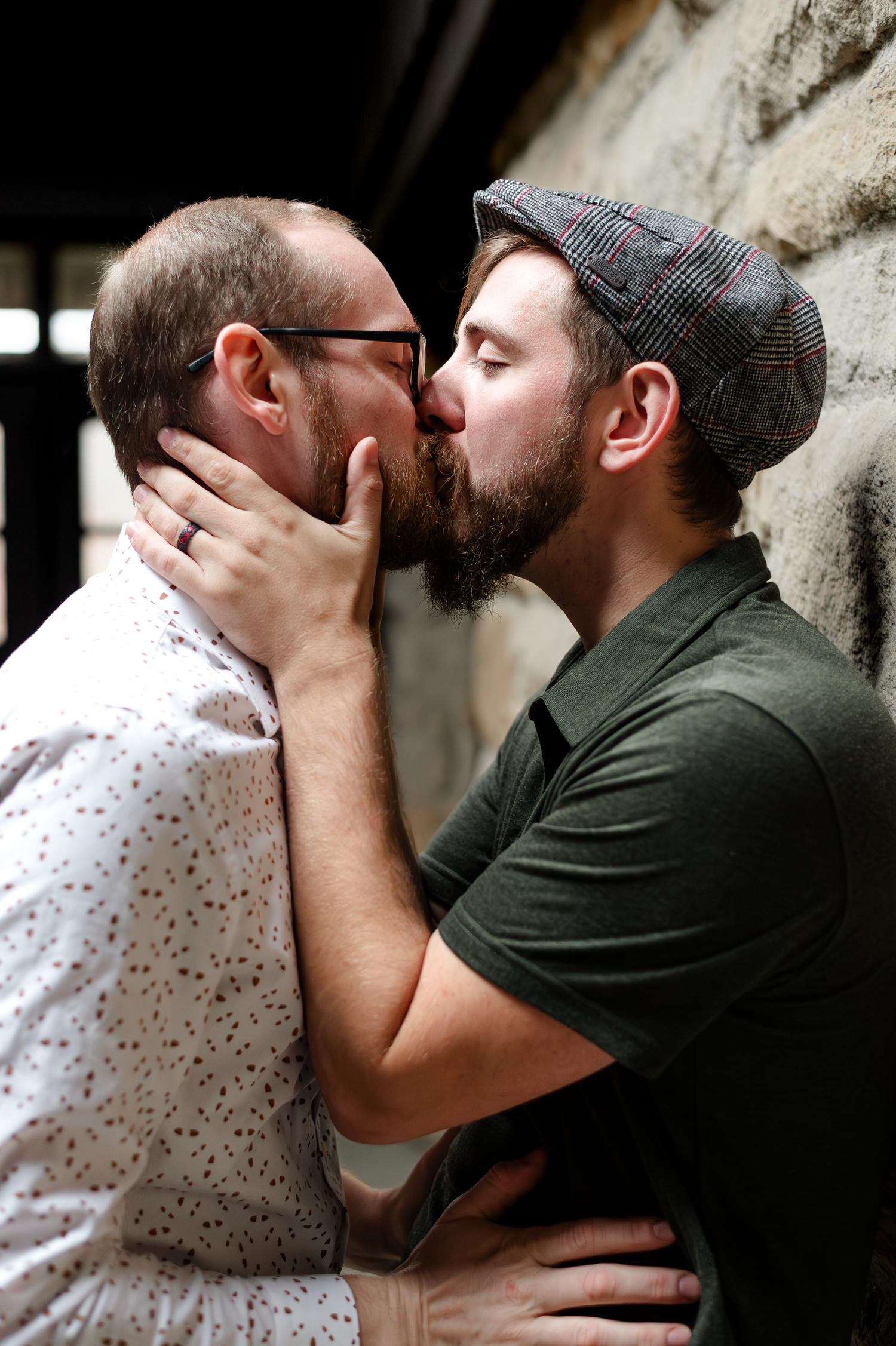 Two bearded men sharing a romantic kiss outdoors, one wearing glasses and a patterned shirt, the other in a cap and green tee.