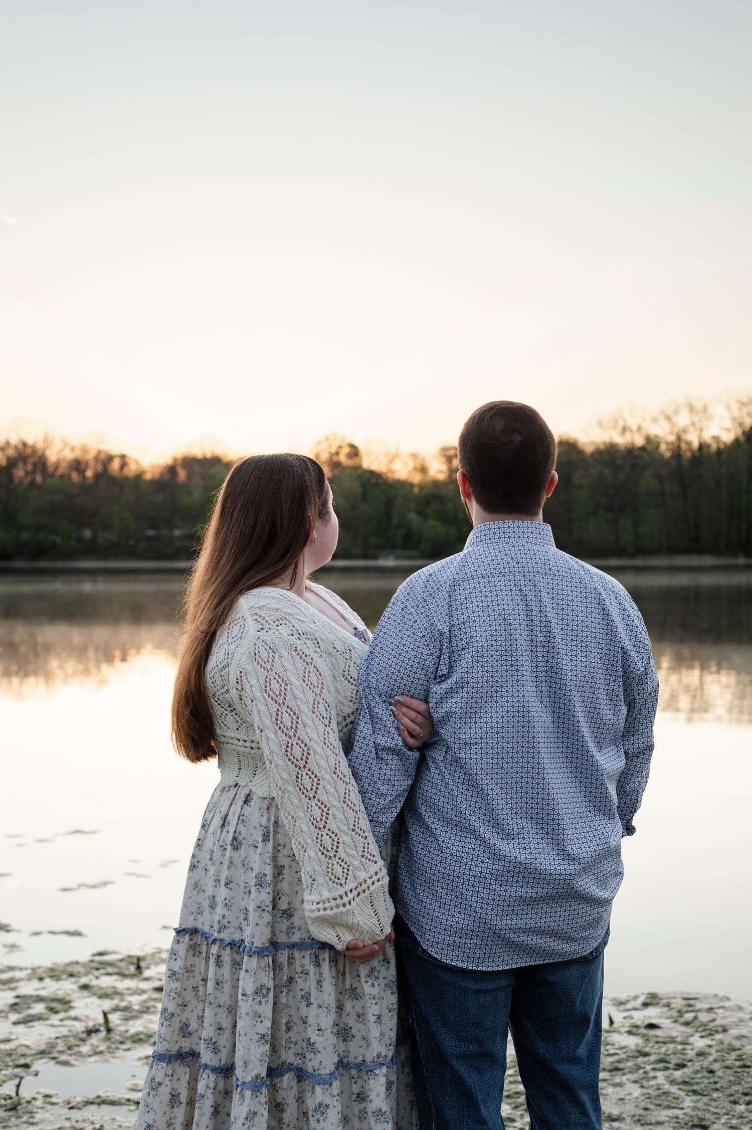 Couple standing together by a calm lake at sunset, woman in floral dress and man in blue shirt, viewed from behind.