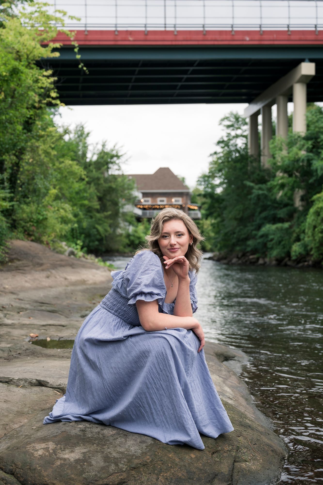 Person in blue dress poses on rocks by a river under a bridge.