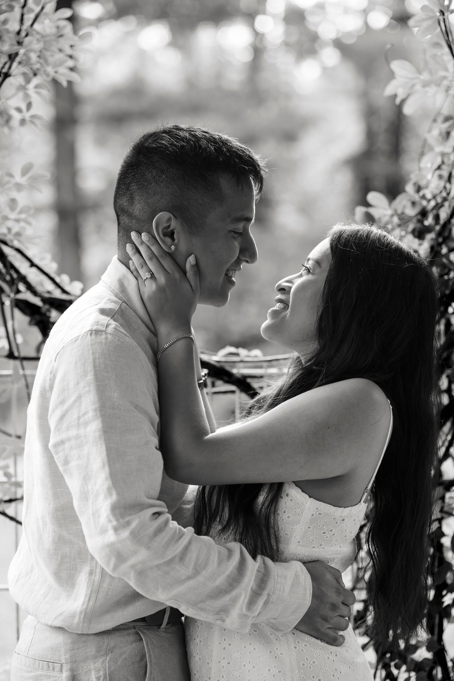 Black and white photo of a couple embracing and smiling at each other outdoors among lush greenery.