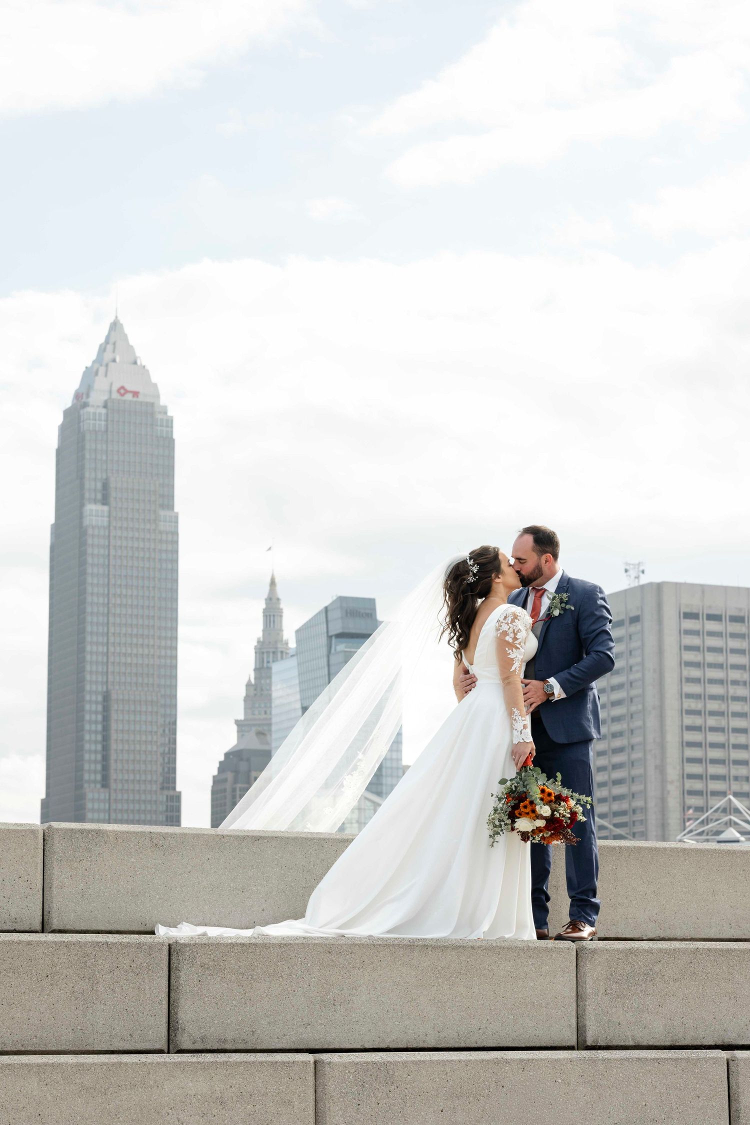 A romantic wedding couple shares a kiss on a rooftop terrace with the Cleveland skyline in the background.