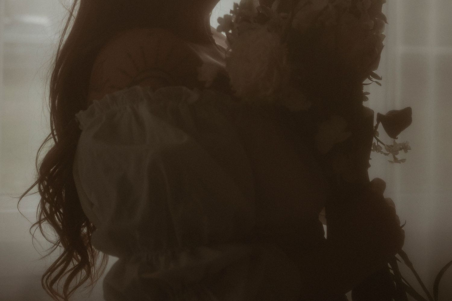 A person in a white dress holds dried flowers in a moody, dimly lit, atmospheric portrait.