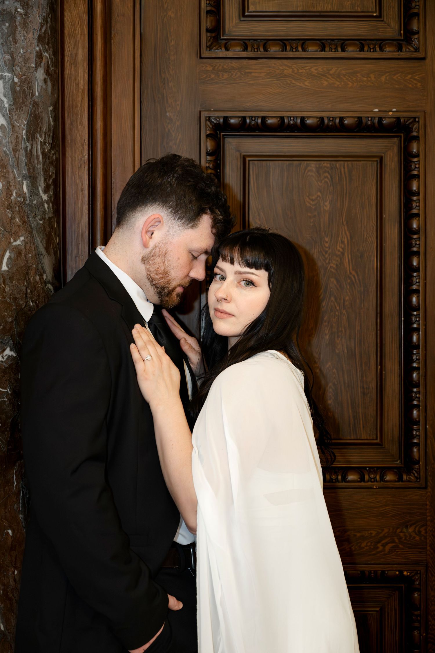 Elegant couple in formal attire embracing near ornate wooden door, man in black suit, woman in white dress.