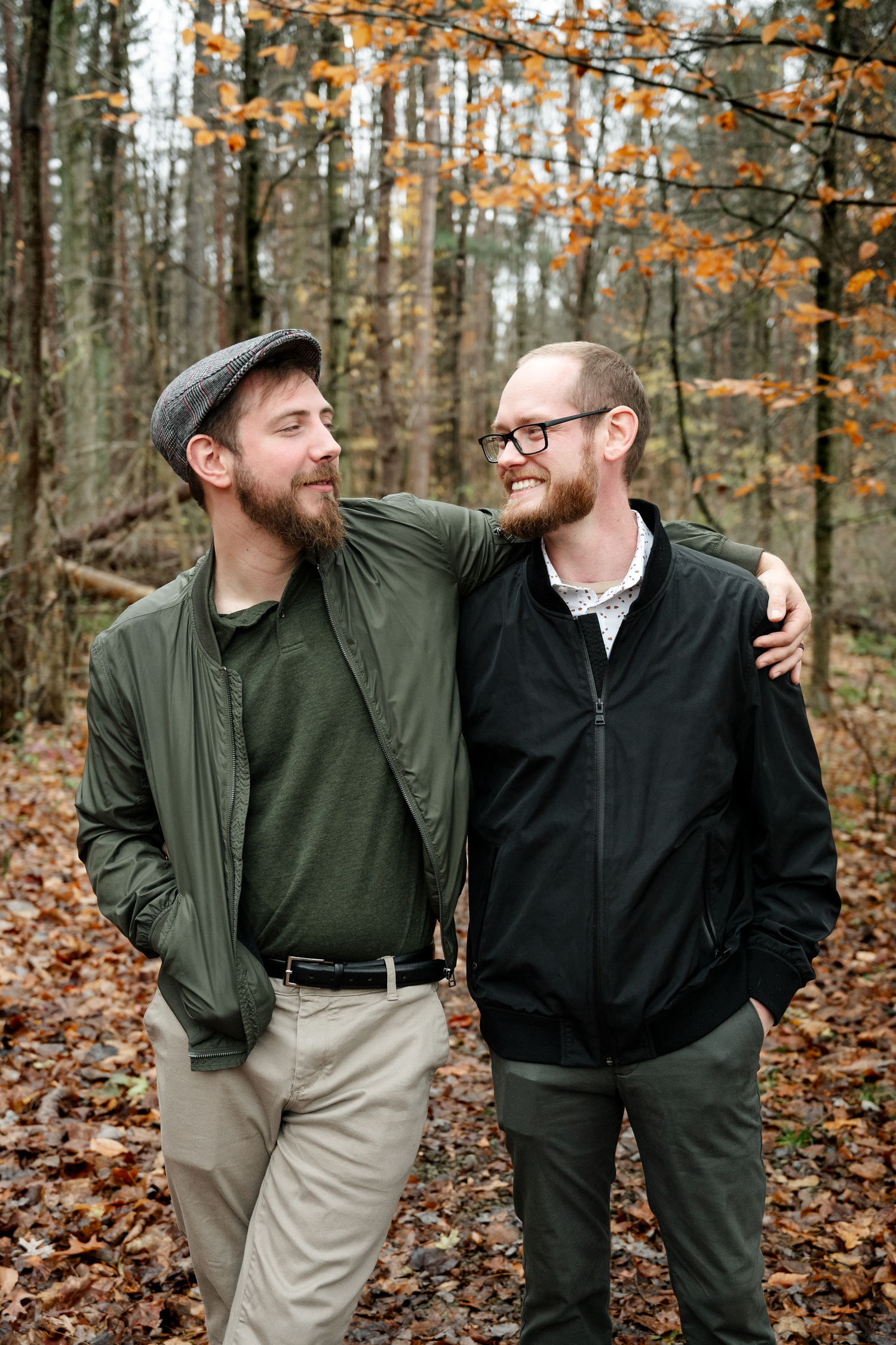 Two bearded men smiling and embracing in an autumn forest, wearing casual jackets and standing among fallen leaves.
