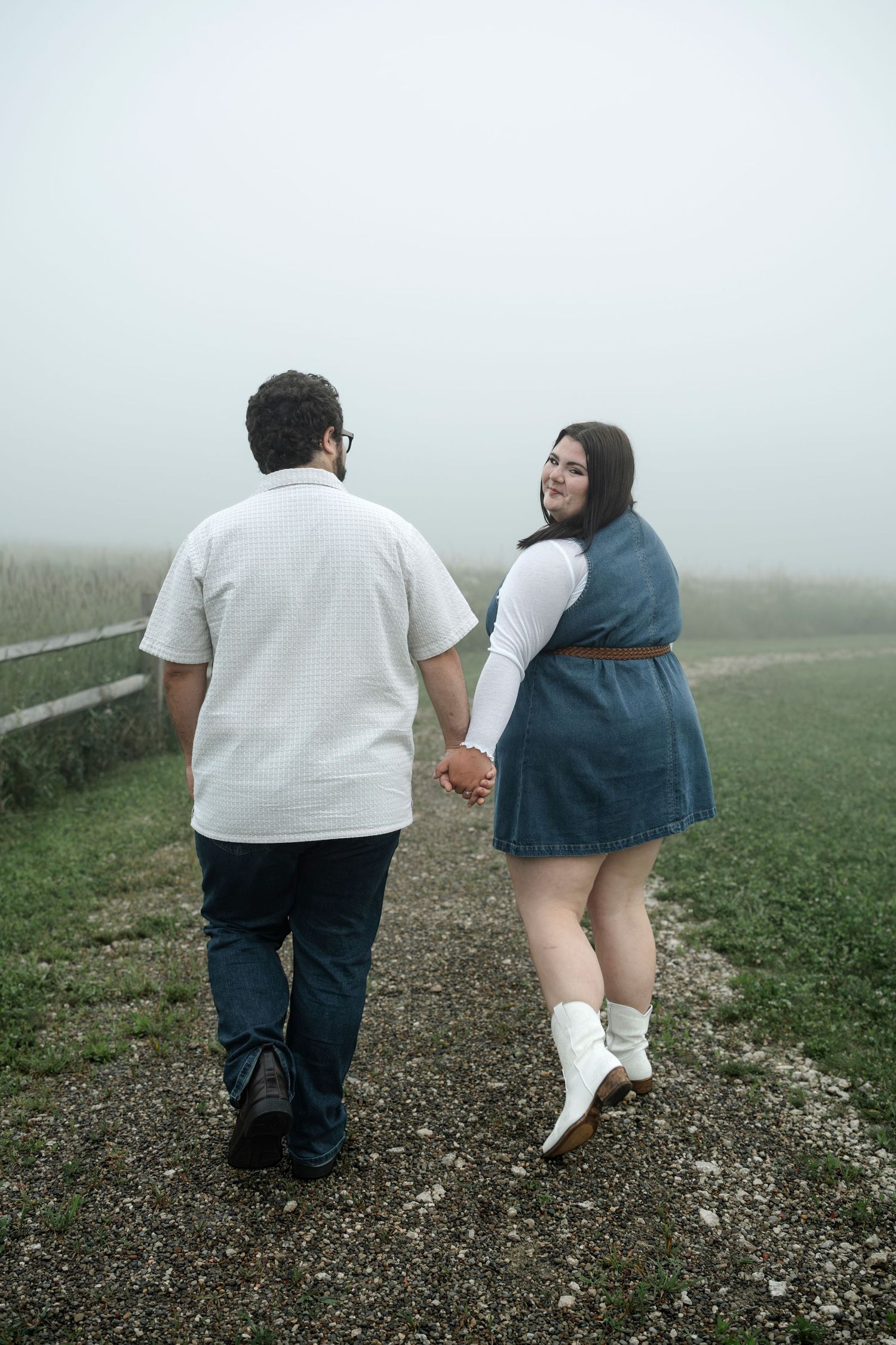 Couple holding hands walking on a foggy path, woman in denim dress and white boots looks back smiling.