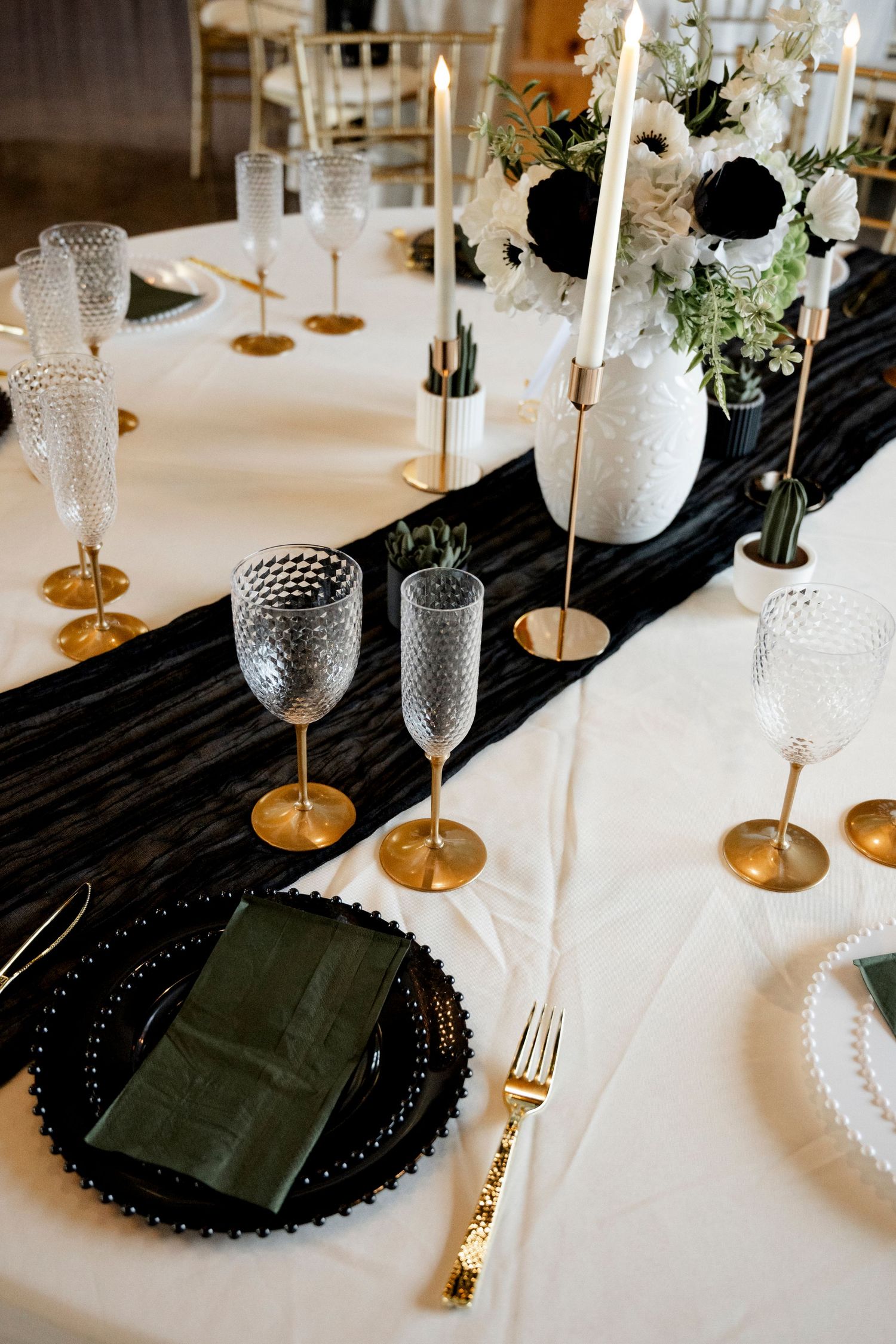 Elegant black and gold wedding table setting with crystal glasses, taper candles, and white floral centerpiece on white linen.