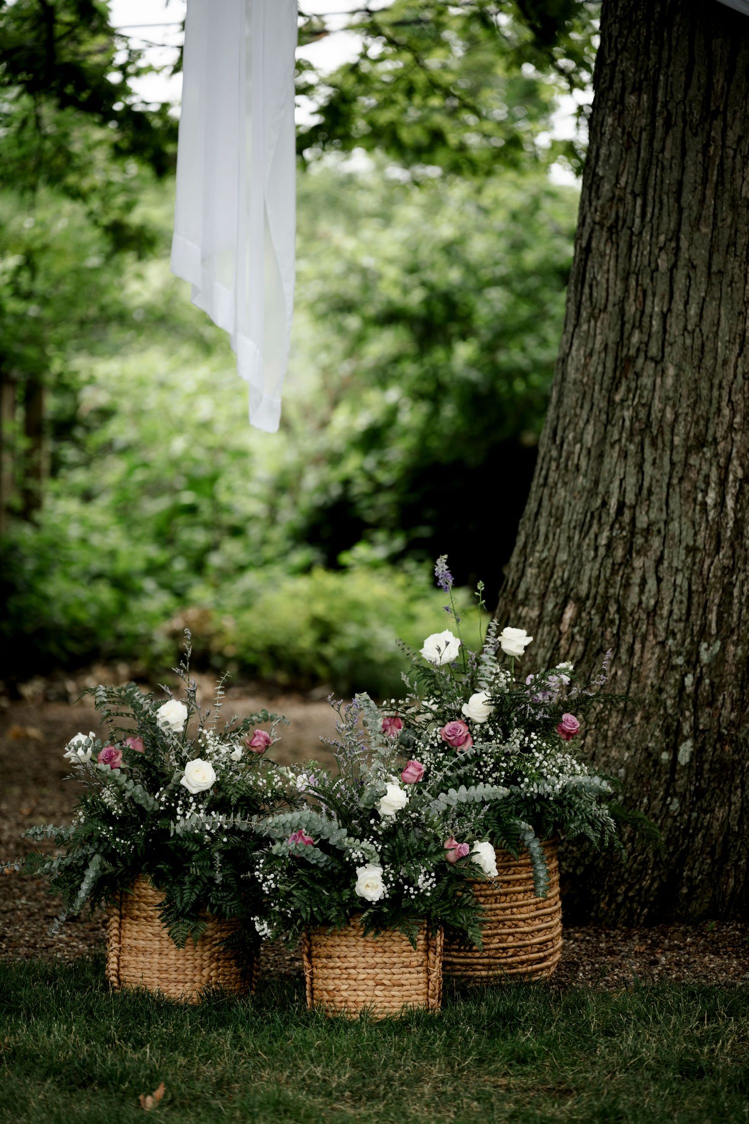Three wicker baskets filled with lush floral arrangements of white and pink flowers sit on grass near a large tree outdoors.