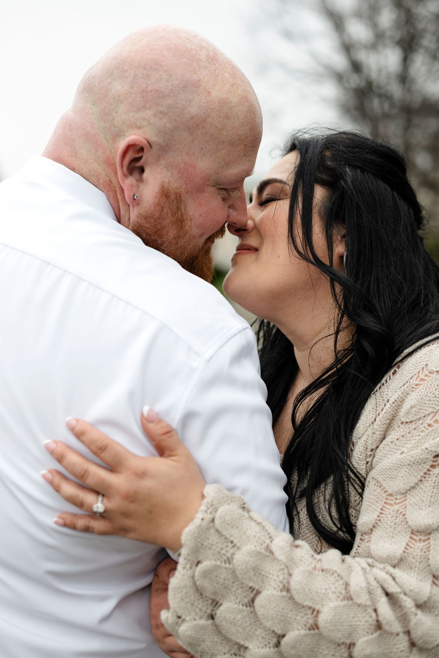 A bald man with a red beard and a dark-haired woman share a romantic nose-to-nose moment, she wears an engagement ring.