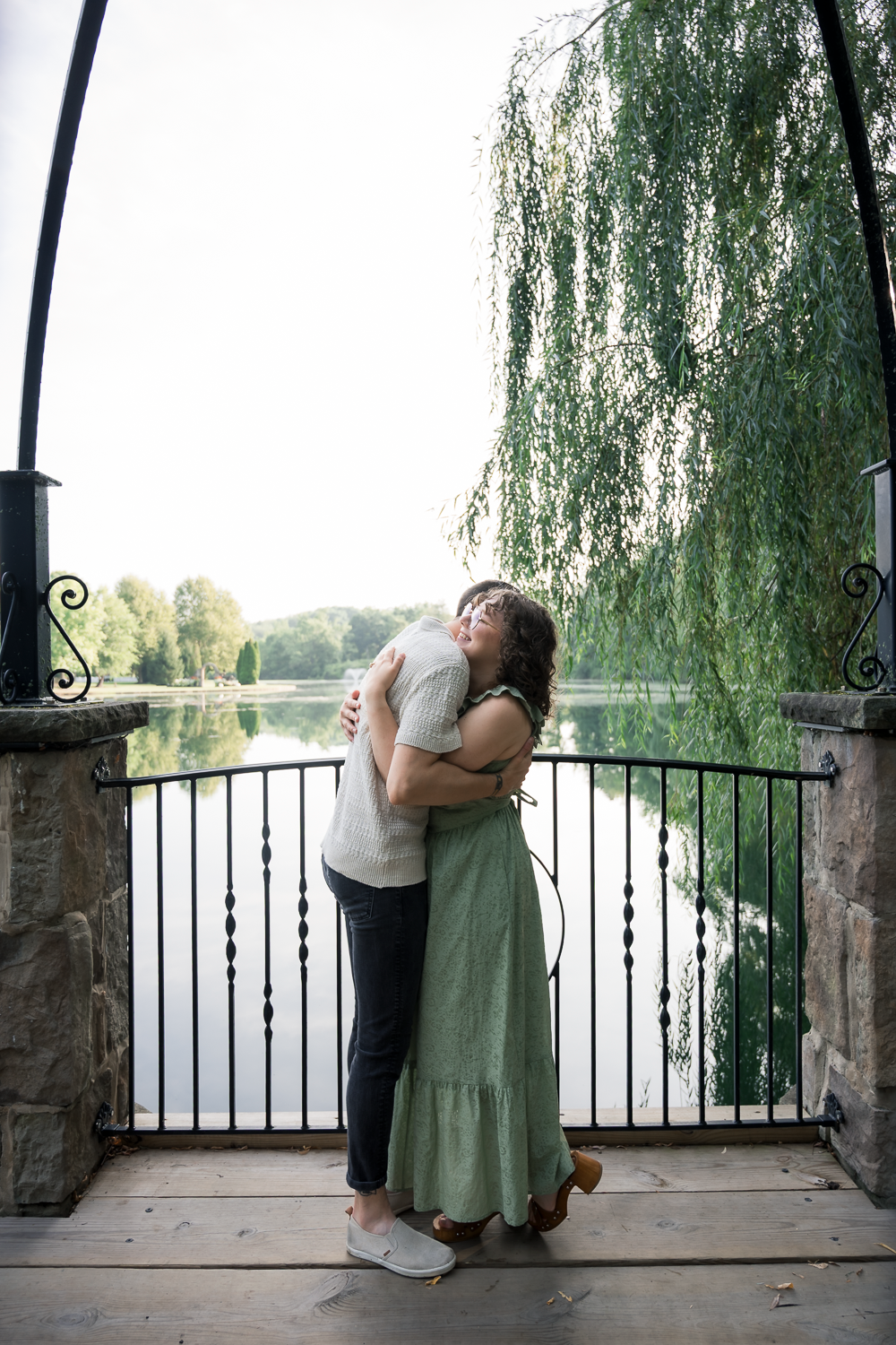 Couple sharing a tender hug on a stone bridge overlooking a serene lake with a weeping willow.
