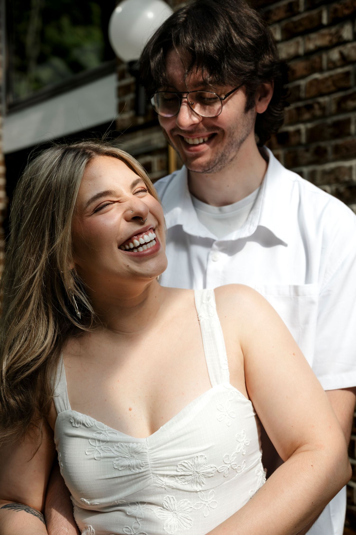 A laughing young woman in a white dress enjoys a joyful moment with a smiling man in glasses outdoors.