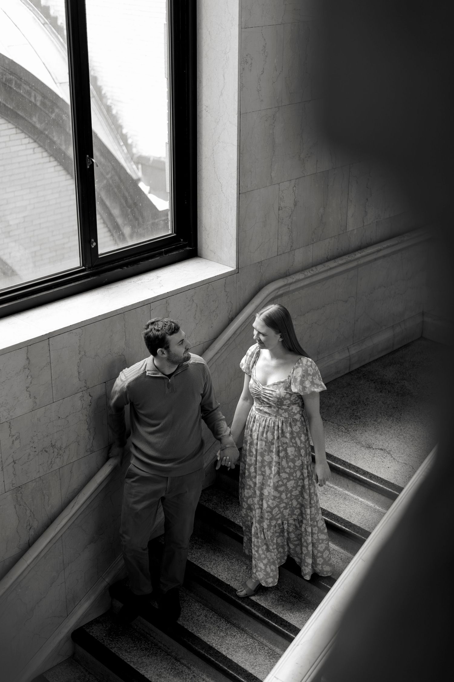 Black and white photo of a couple holding hands on elegant stone stairs, gazing at each other near a large window.