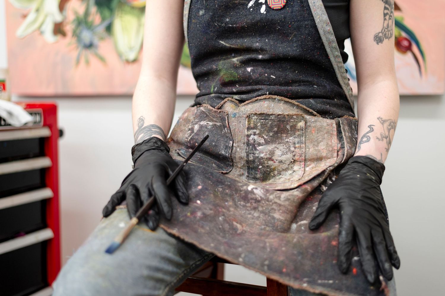 Close-up of a tattoo artist wearing black gloves and paint-splattered work clothes in their studio.