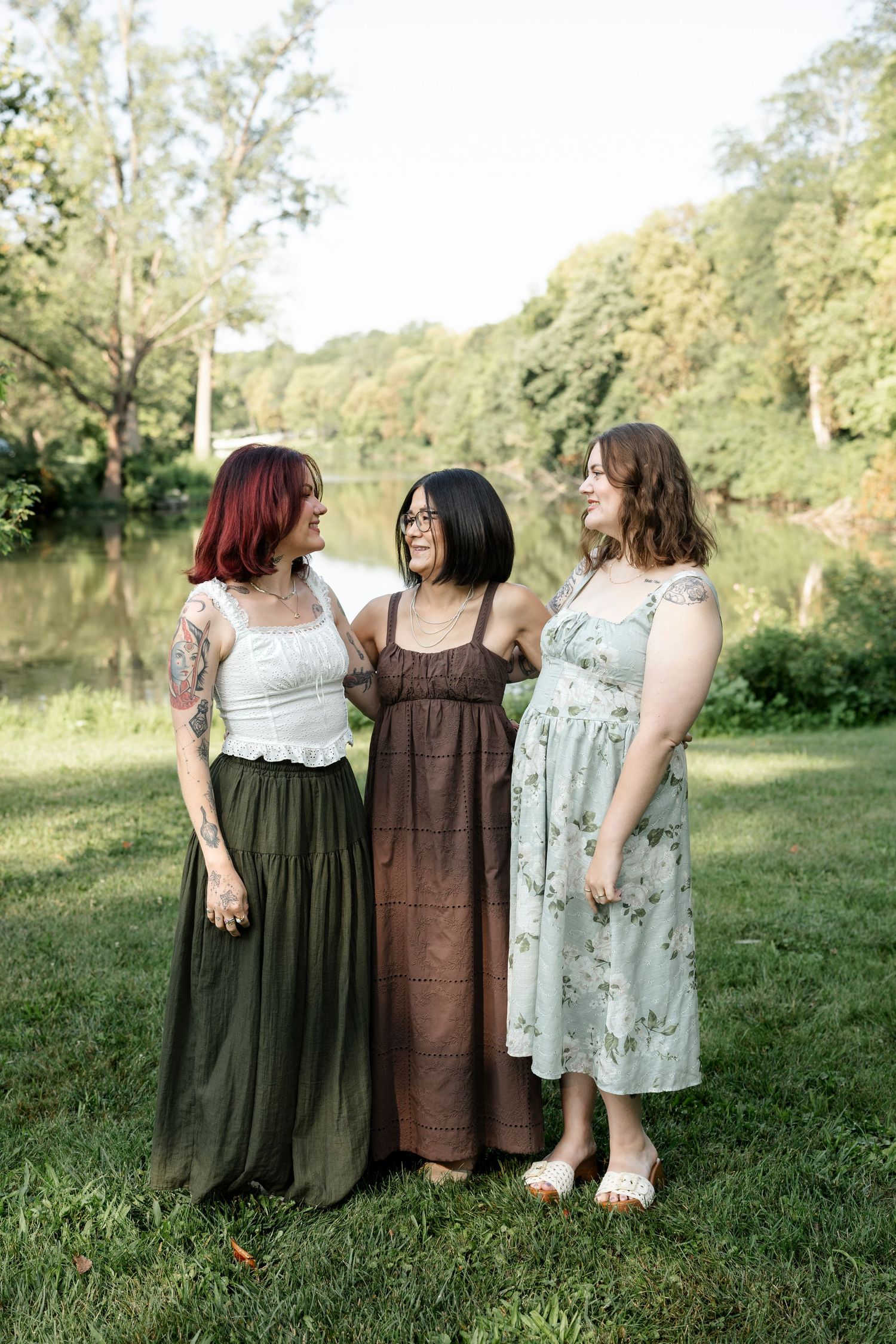 Three women in summer dresses smiling together outdoors near a river surrounded by lush green trees.