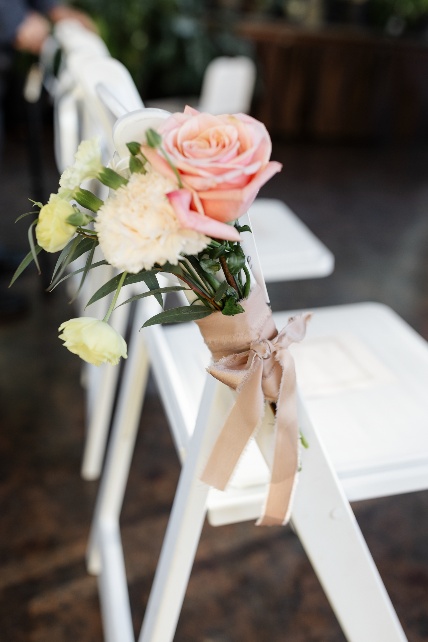 A peach rose and carnation bouquet tied with a beige ribbon decorates a white folding chair at a wedding ceremony.