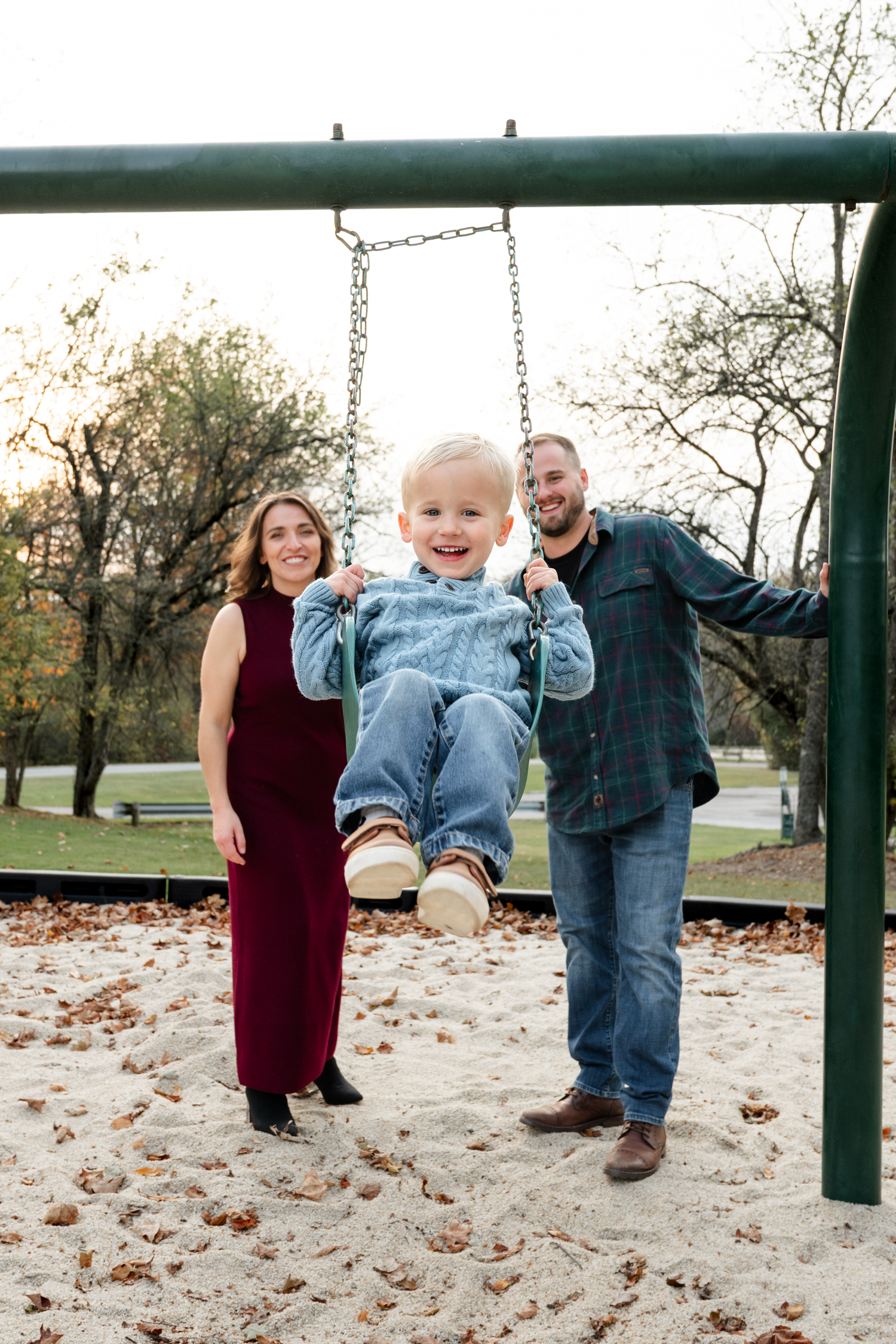 Family enjoying time together pushing a child on a swing set in a park with fallen leaves.