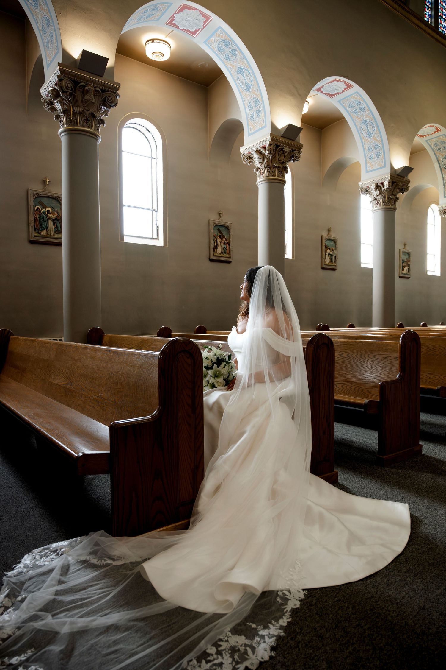 Bride in white gown and veil sits in church pew, long train flowing down aisle between ornate columns.