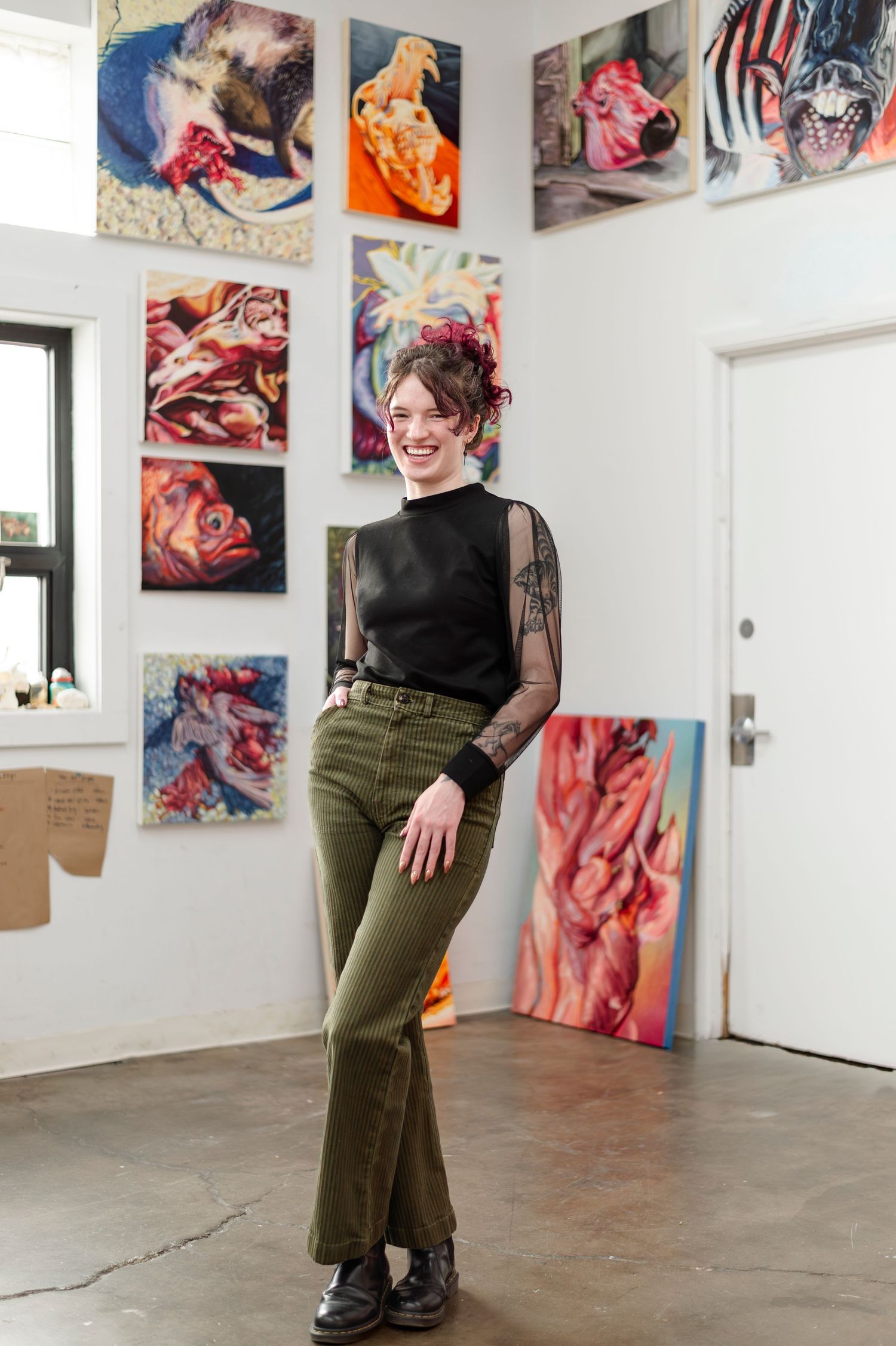 Artist stands in art studio with colorful abstract paintings displayed on walls in casual attire.