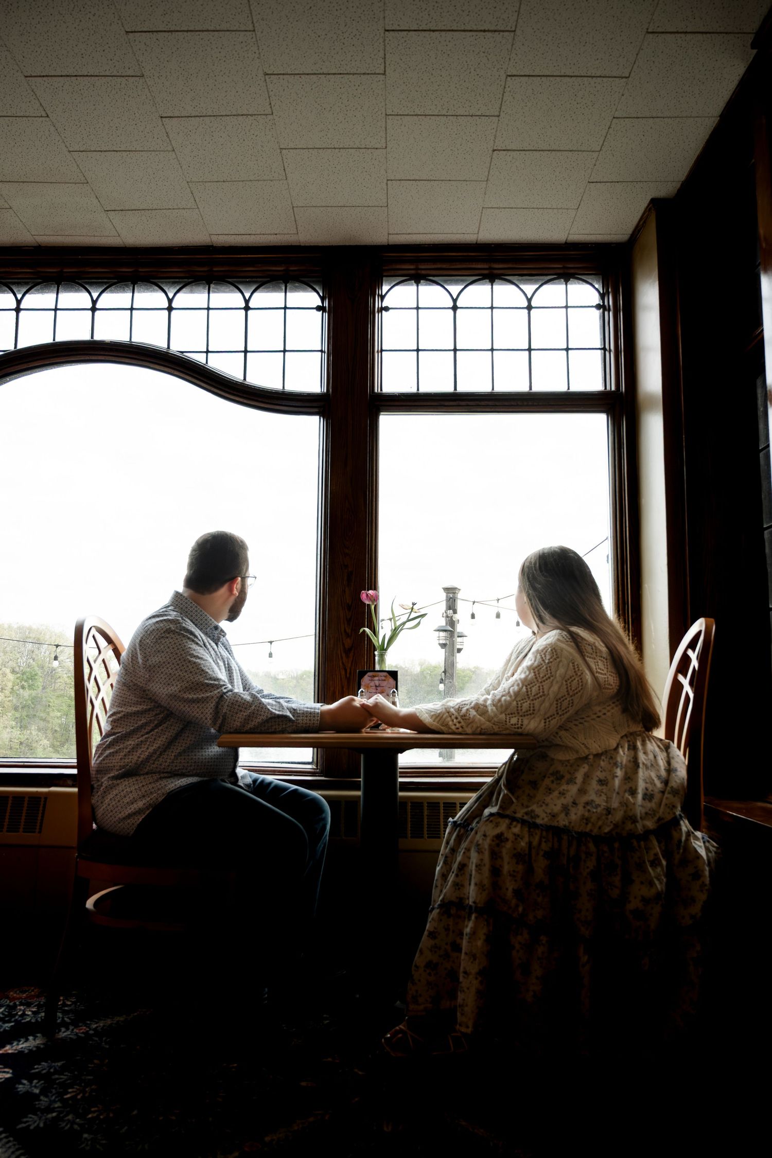Couple holding hands at a restaurant table by large arched windows, sharing a romantic moment with a pink flower centerpiece.