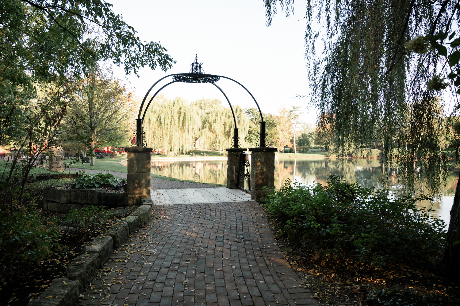 A scenic stone path winds through a park with weeping willow trees and autumn leaves near a tranquil pond.