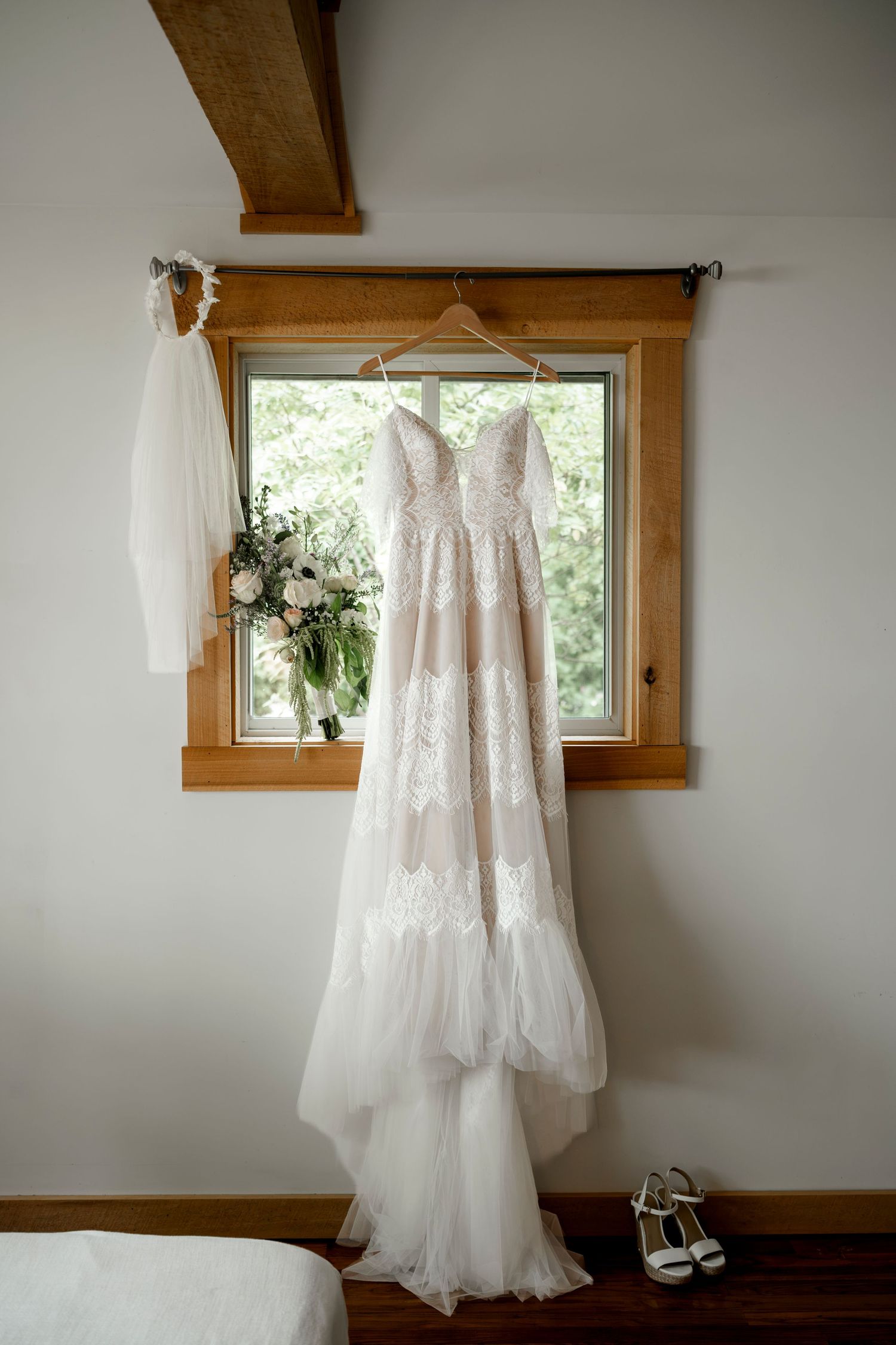 Elegant lace wedding dress hanging by a window with bridal bouquet and heels displayed in a rustic venue.