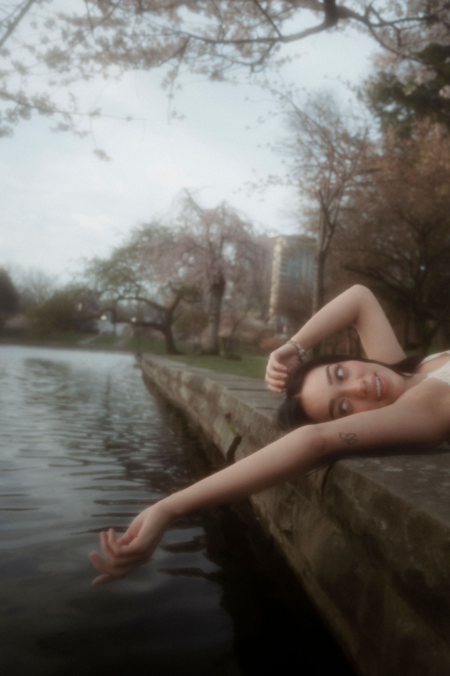 Young woman lying on a stone ledge by a tranquil river, surrounded by cherry blossom trees in soft focus.