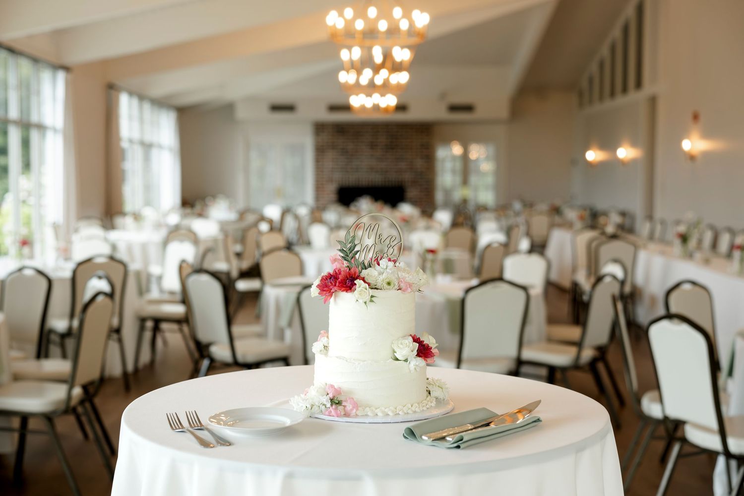 Elegant two-tier white wedding cake with pink florals on a draped table in a bright reception hall.