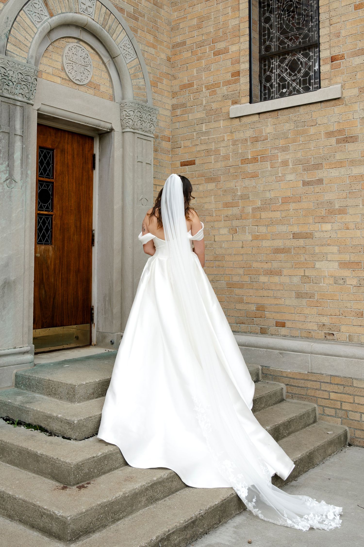 Bride in white off-shoulder gown with long veil stands on stone church steps, viewed from behind.