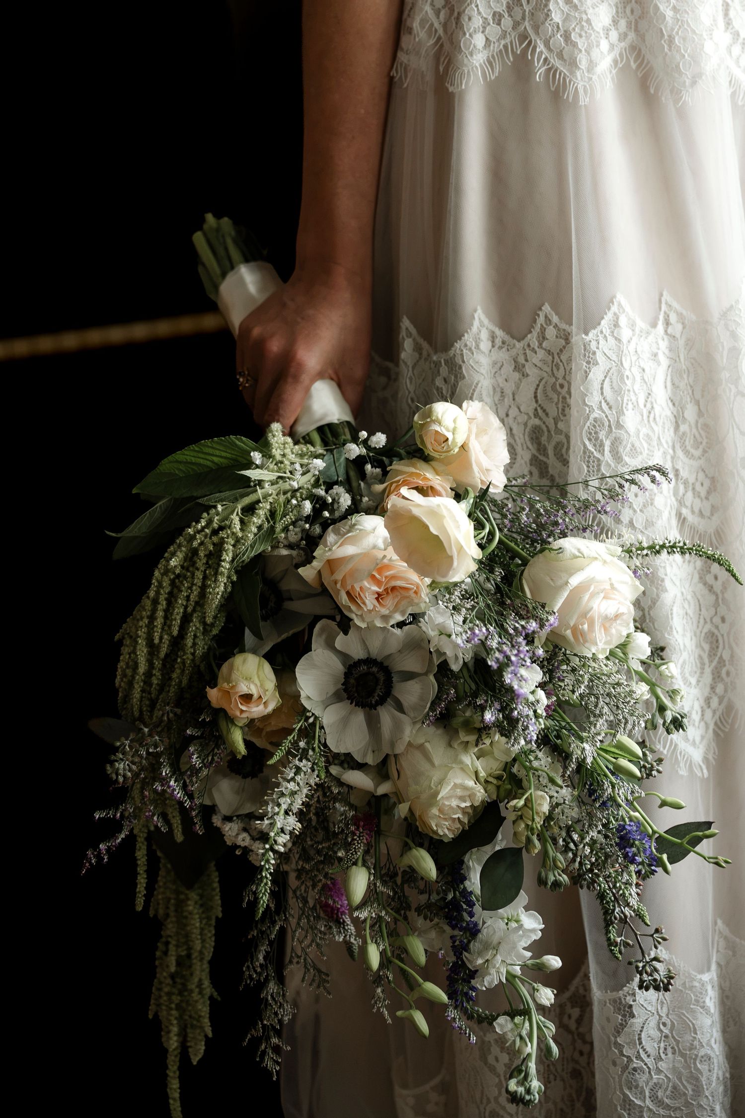 Bride in lace wedding dress holding elegant bouquet of white roses, anemones, and wildflowers.