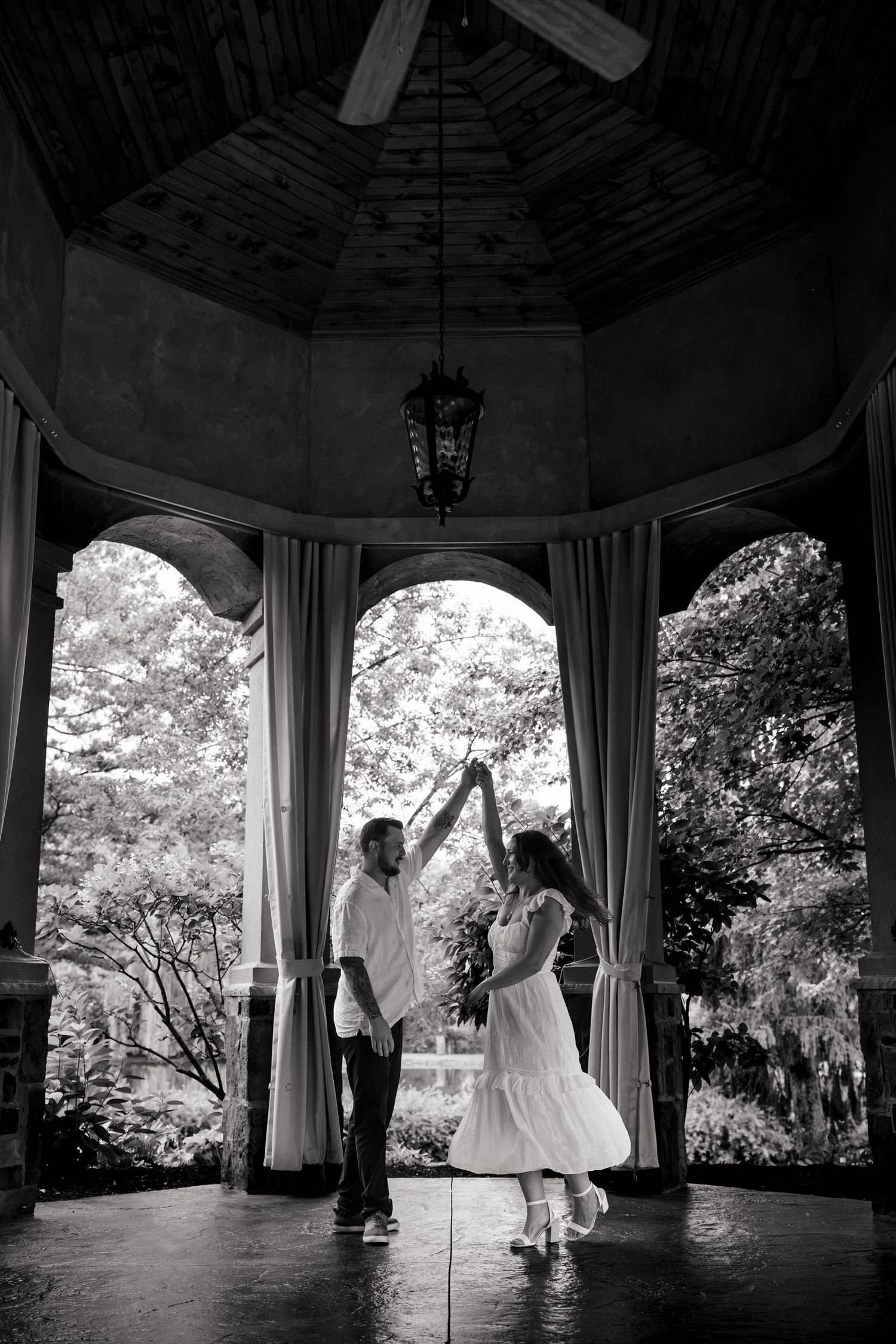 Couple dancing in elegant gazebo with arched columns and hanging lantern, surrounded by lush trees, black and white photo.