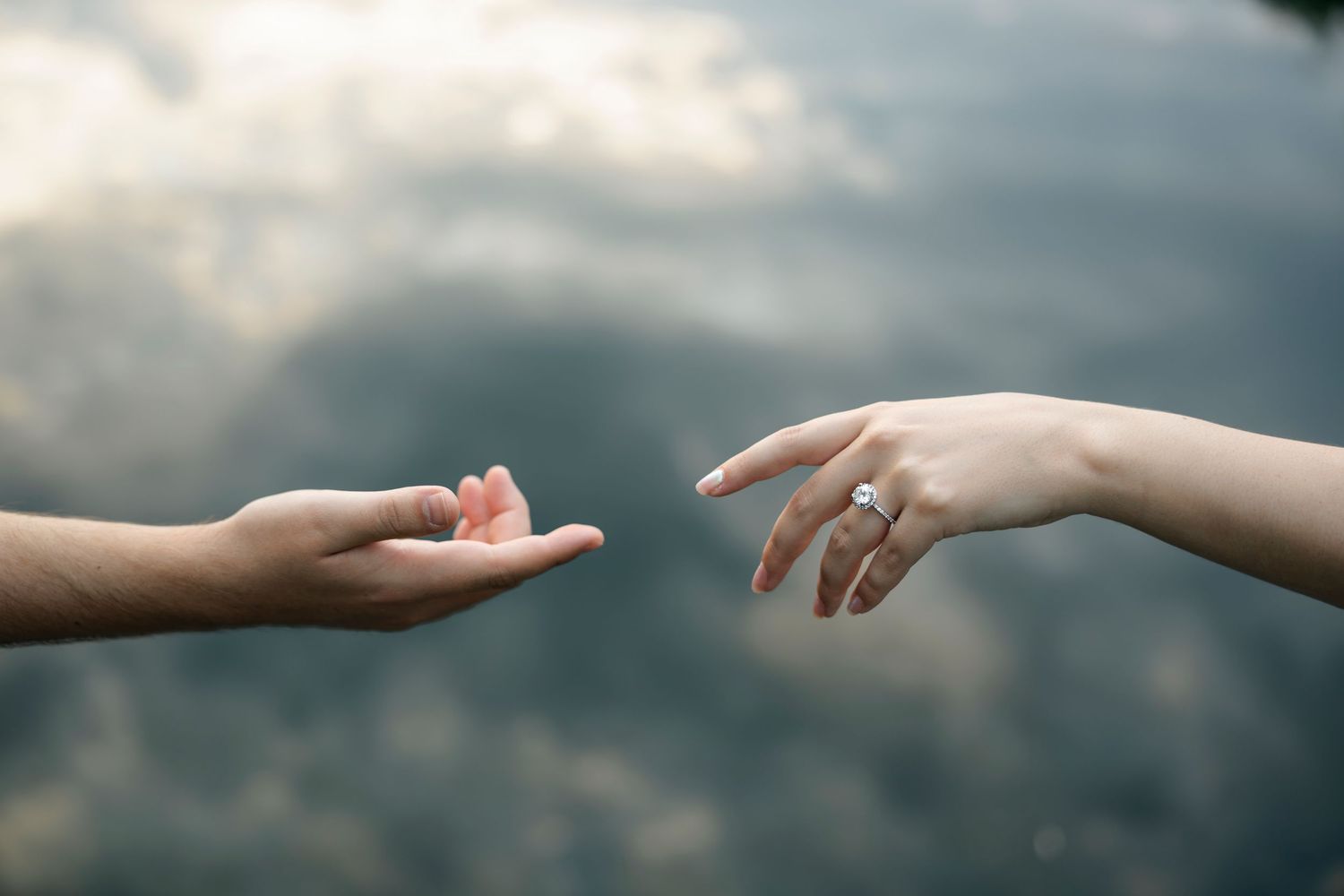 Two hands reaching toward each other, one wearing a diamond engagement ring, against a cloudy sky background.