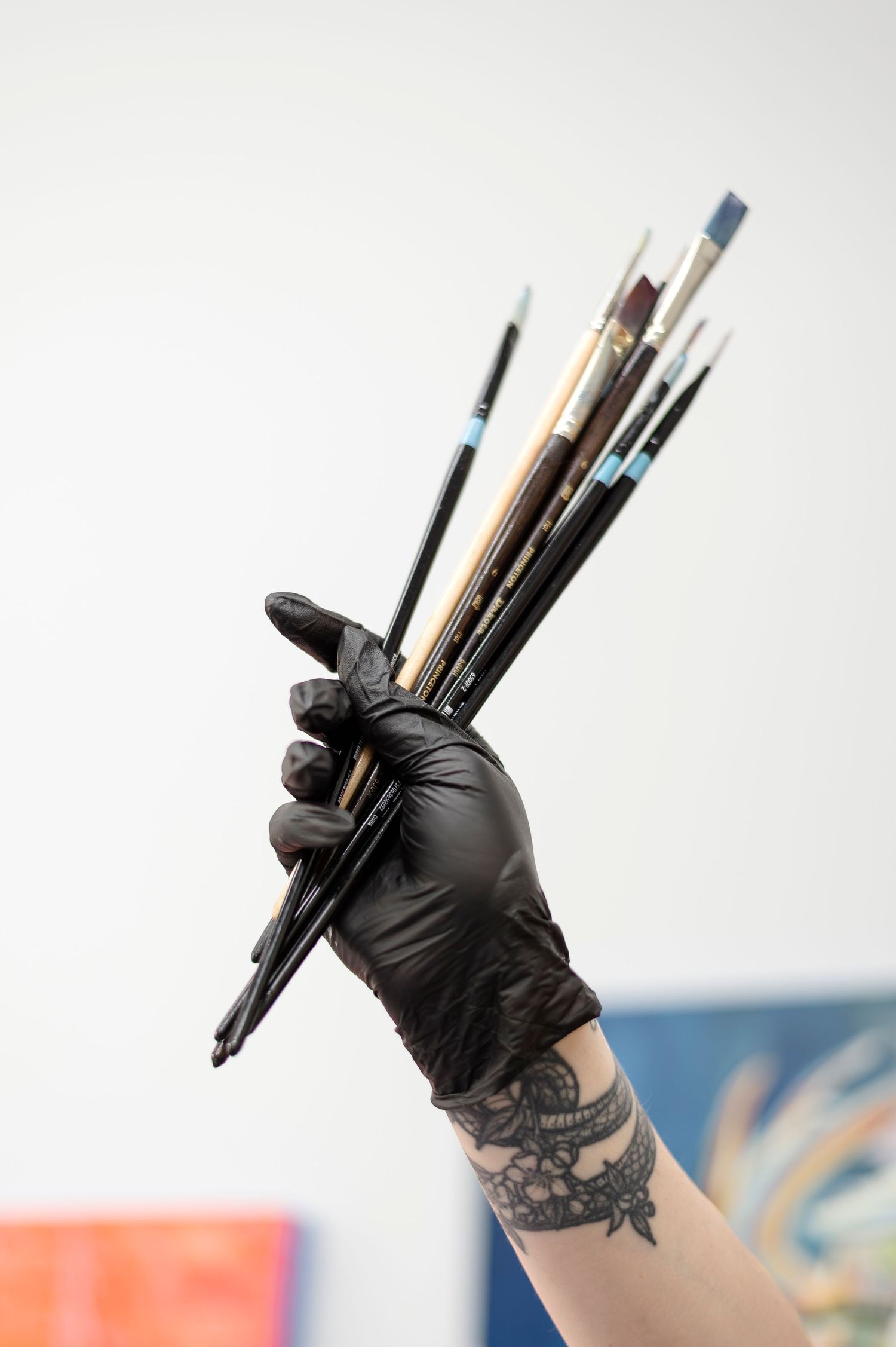 Artist holding paint brushes in black gloves with tattoo visible on wrist against blurred background.