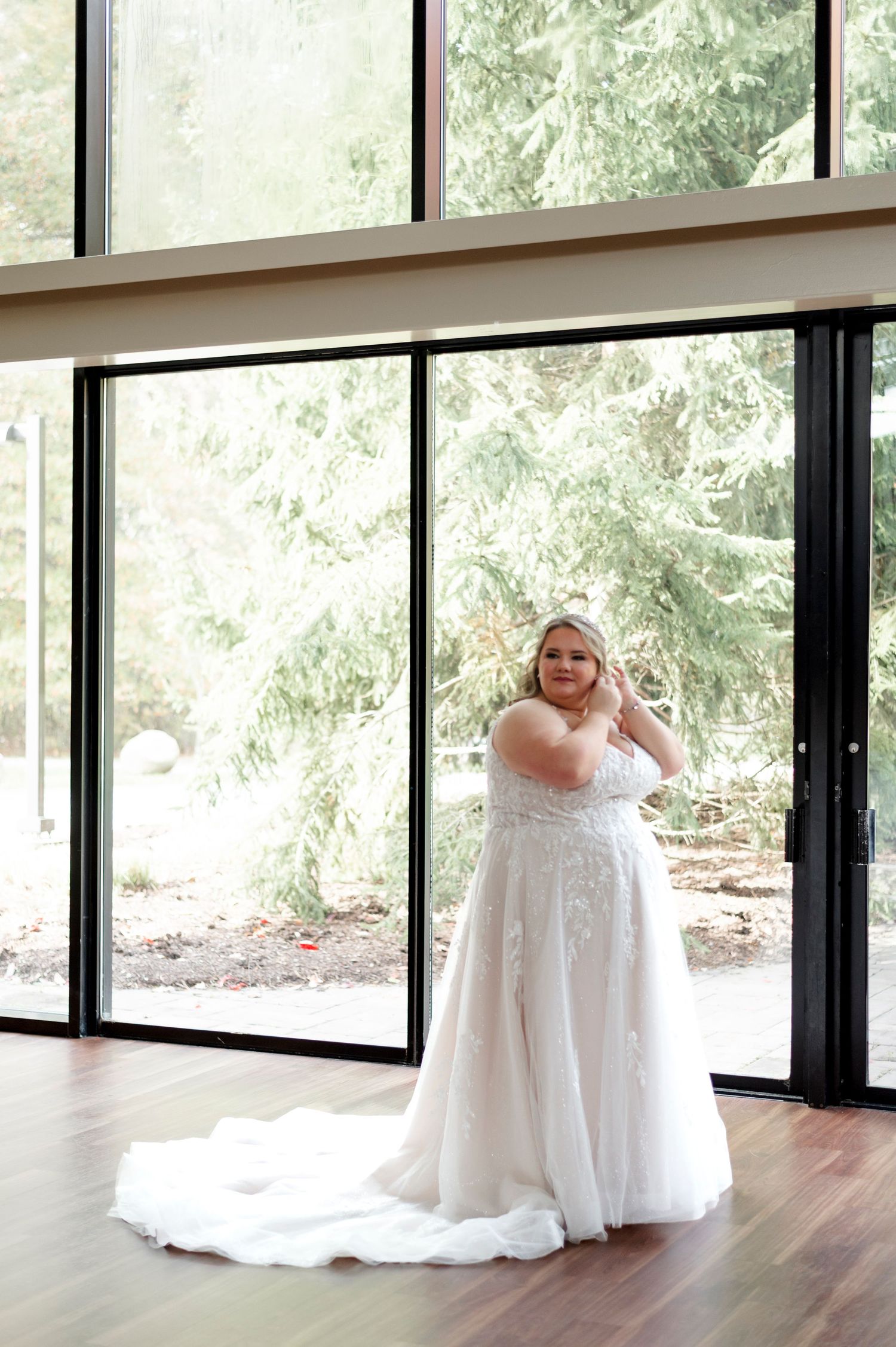 A bride in a flowing white lace gown with train poses elegantly in front of floor-to-ceiling windows with natural light.