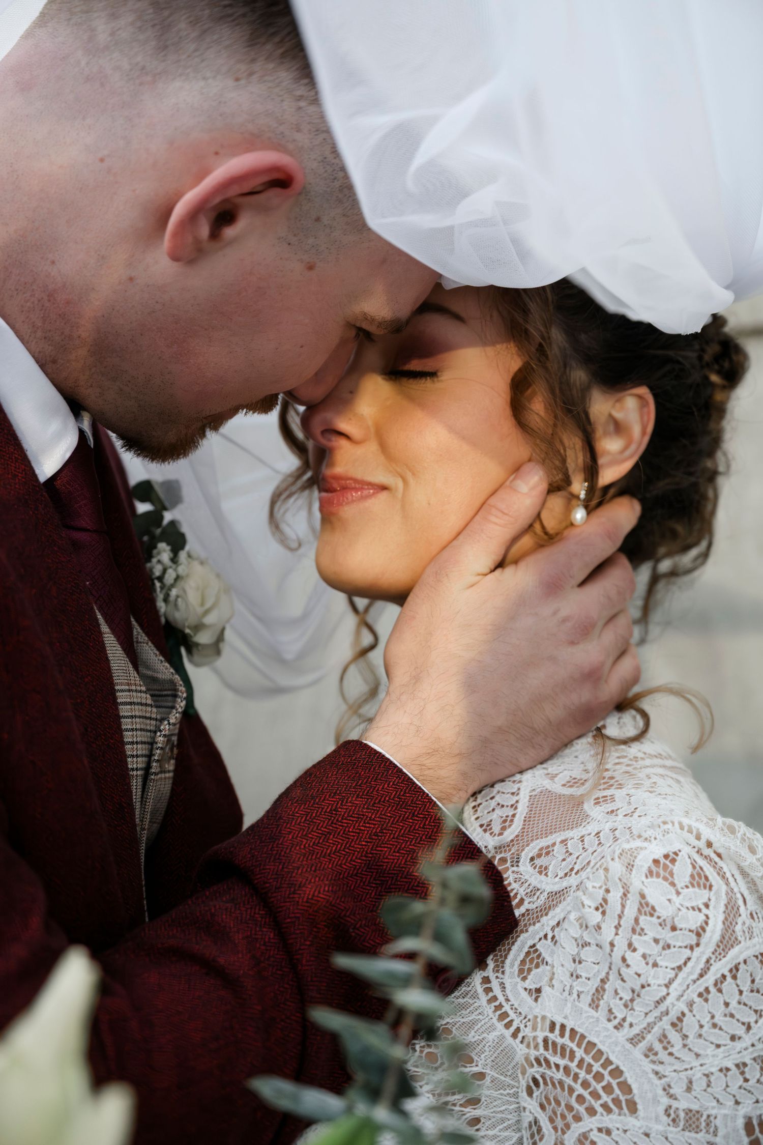 A bride and groom embrace tenderly on their wedding day, with the bride wearing a lace wedding dress and the groom wearing a burgundy suit.