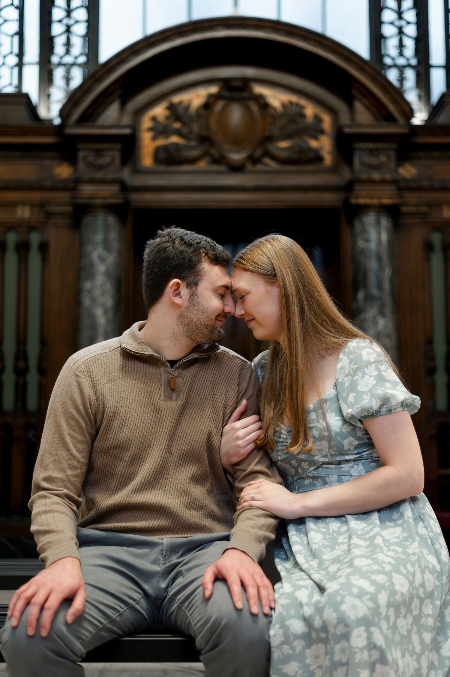 A couple leans foreheads together romantically, seated before an ornate wooden architectural backdrop.